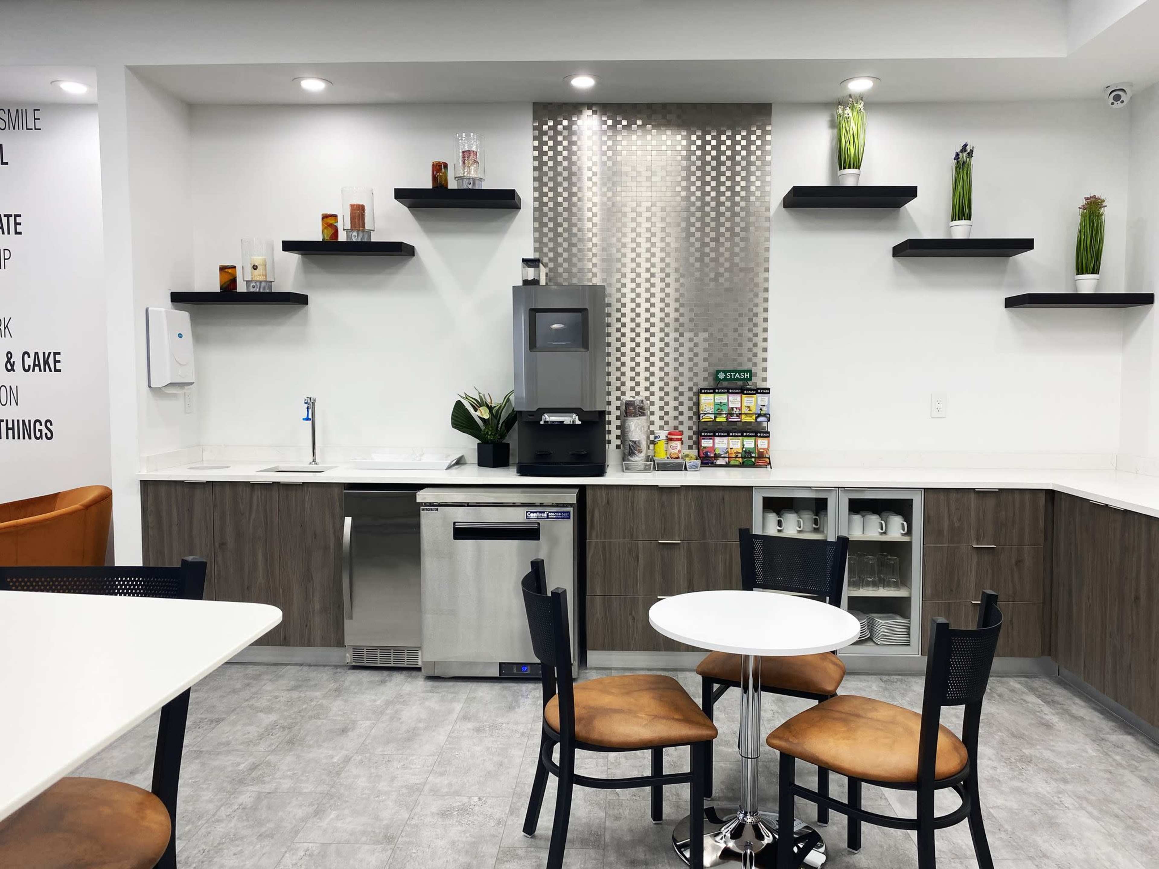 A modern kitchen area features a small round table with chairs, a sink, a refrigerator, shelves with various items, and a coffee station.