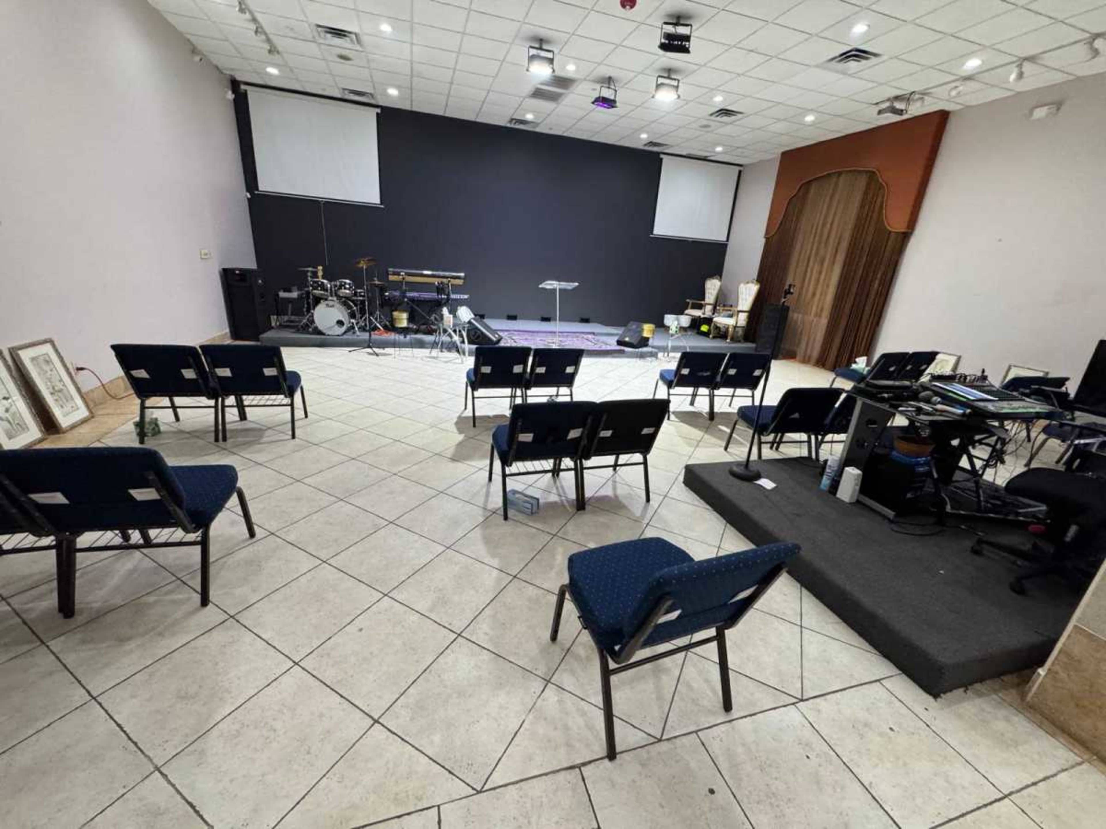 The image shows a spacious room set up for a gathering, featuring rows of blue chairs facing a stage with musical instruments and a sound setup.