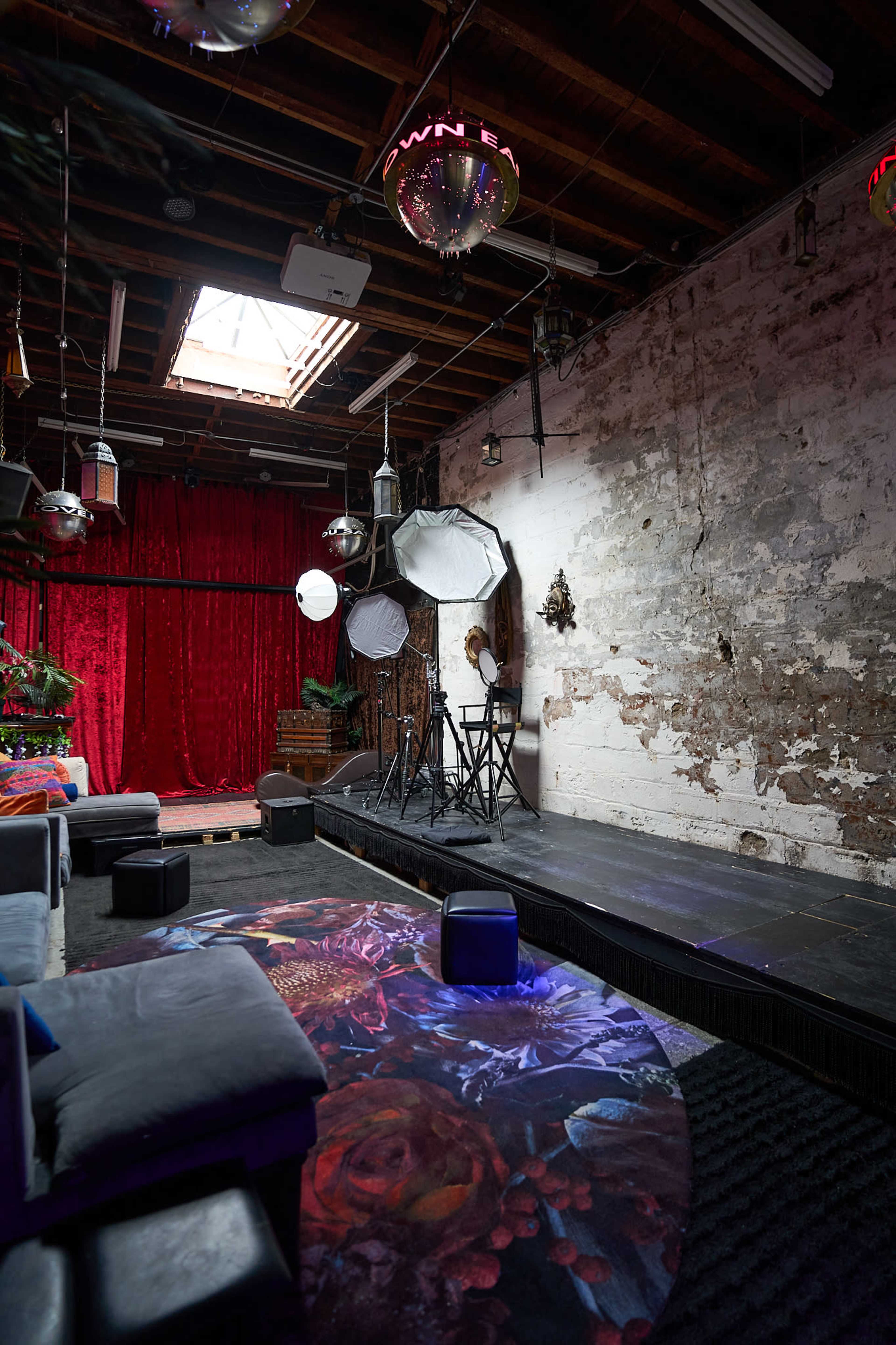 The image shows a dimly lit room with a raised stage, surrounding seating, and a backdrop of red curtains, along with various lighting fixtures.