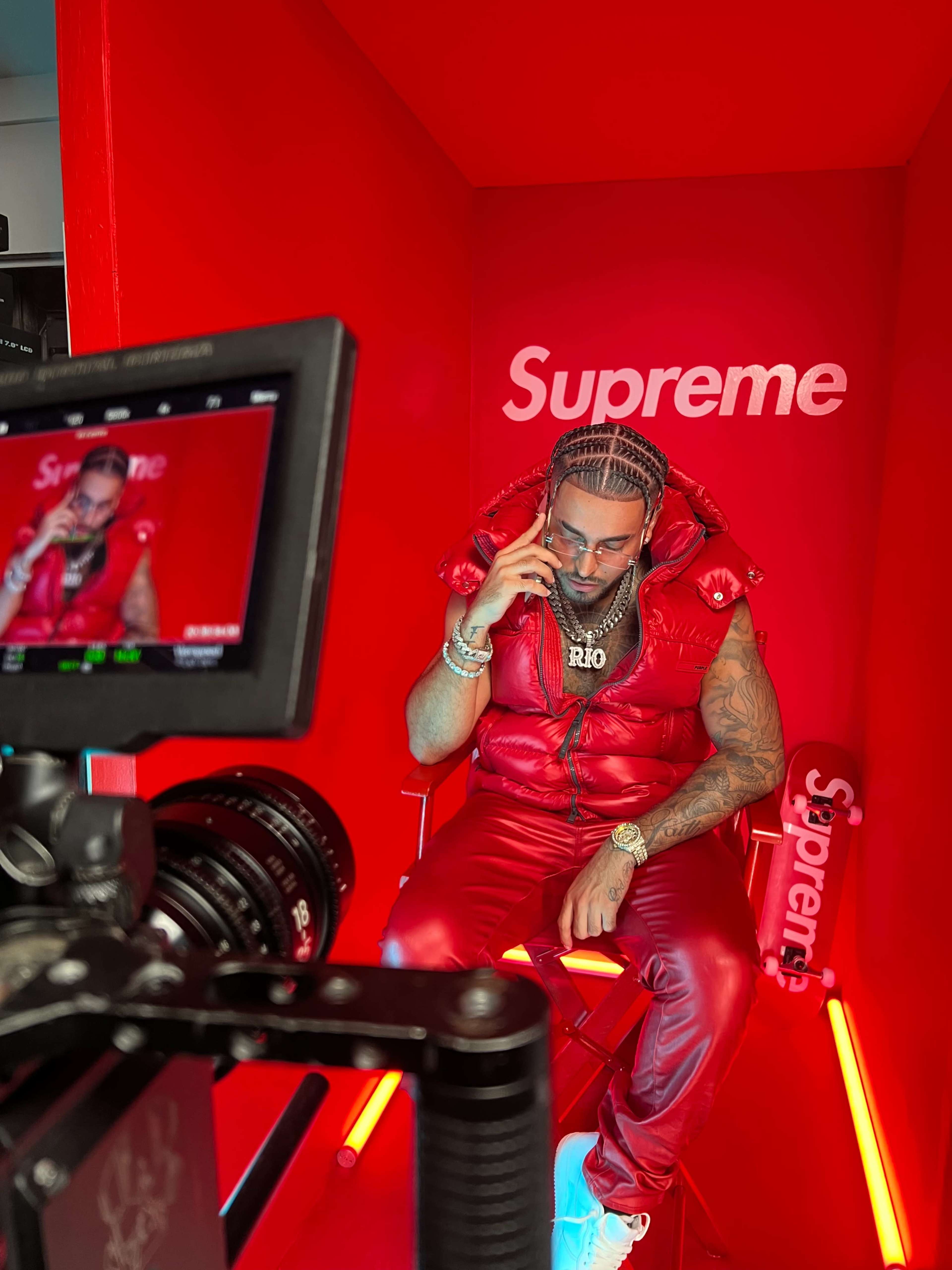 A man wearing a red puffer vest and matching pants sits in a red-lit studio, holding a phone while looking down, with a camera focused on him in the foreground.