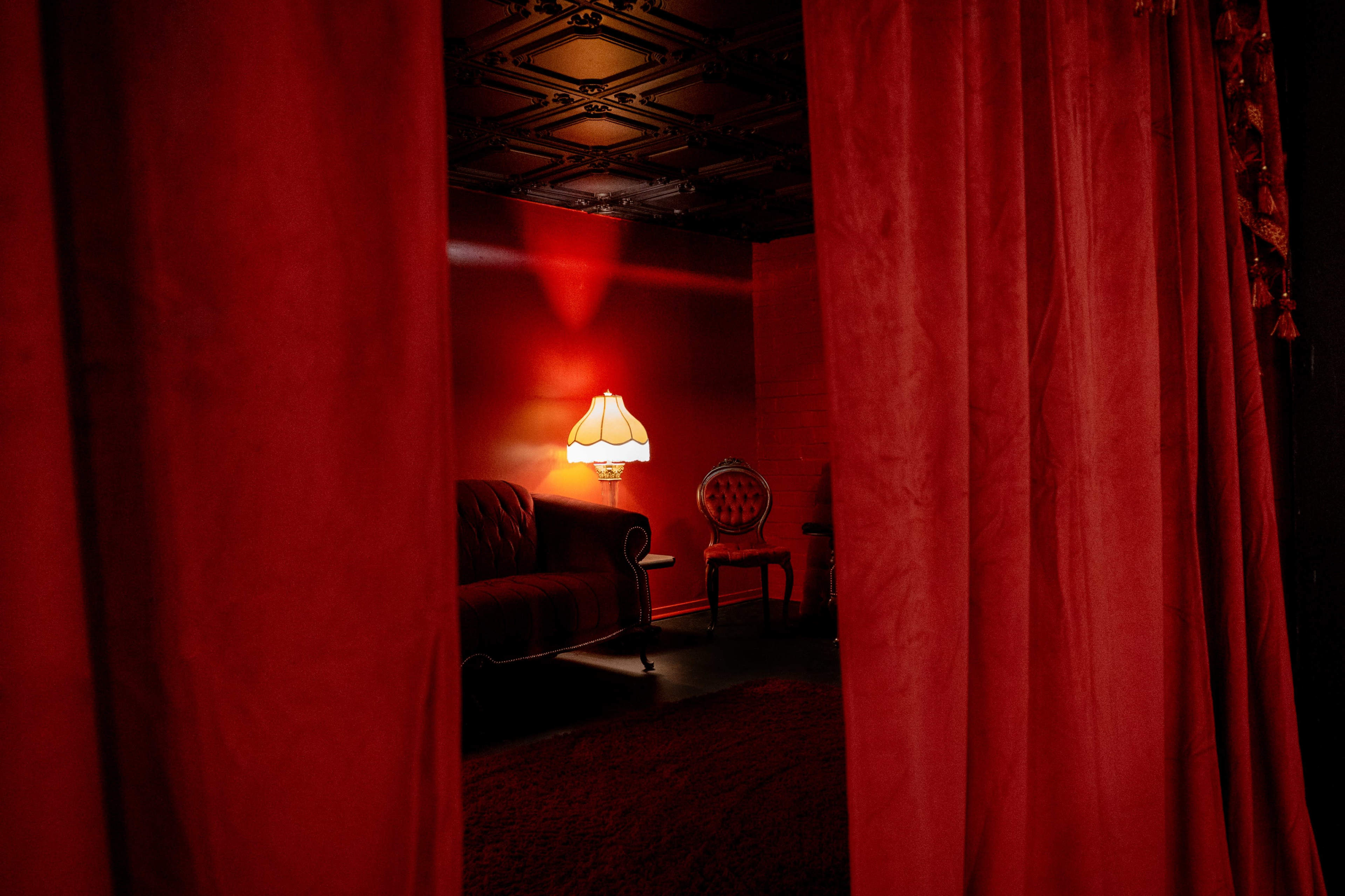 The image shows a dimly lit room with red walls, a plush sofa, and a vintage lamp, framed by red curtains.