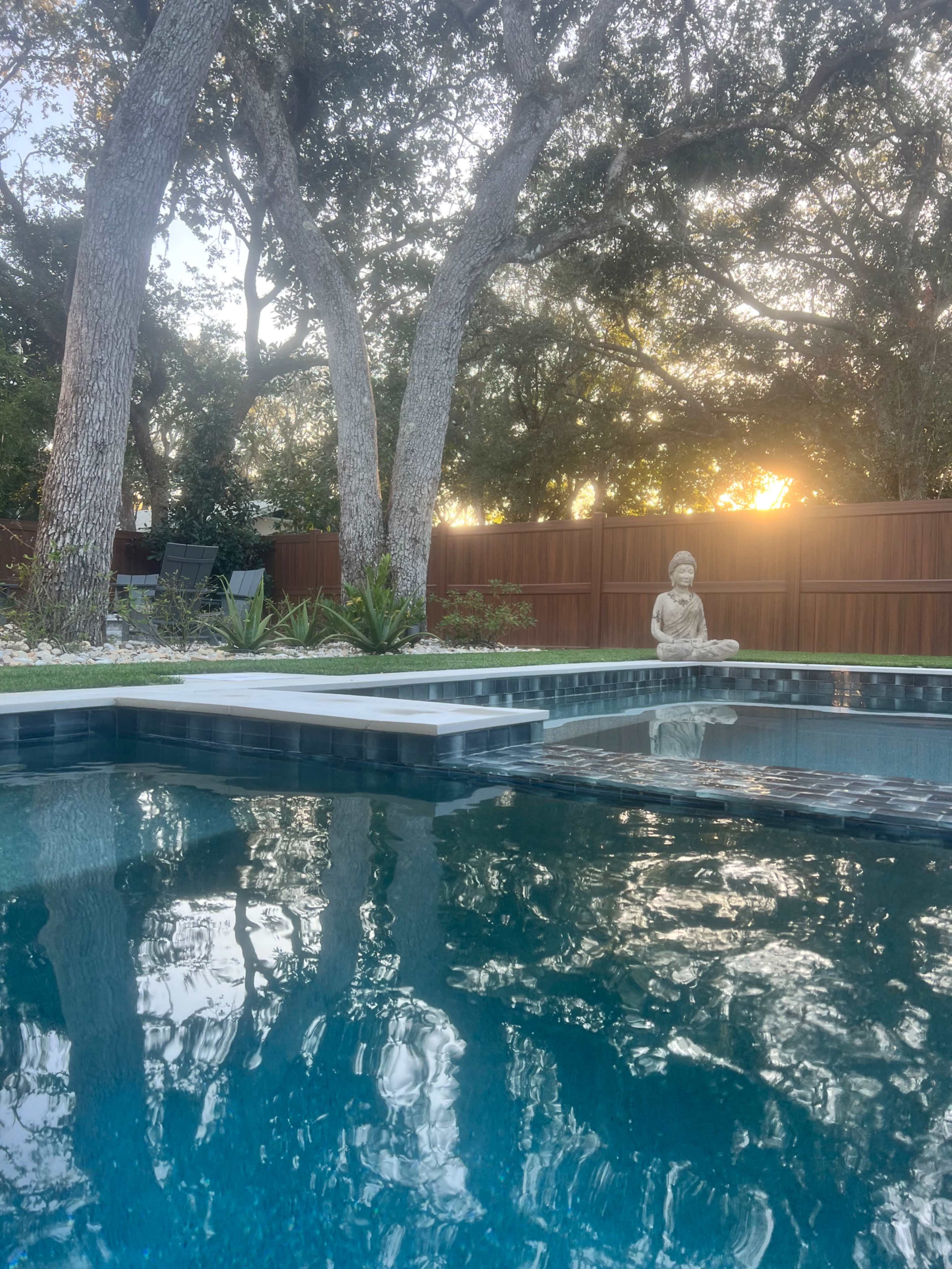 A tranquil pool scene features a statue of a seated figure beside water reflected by surrounding trees and a setting sun.