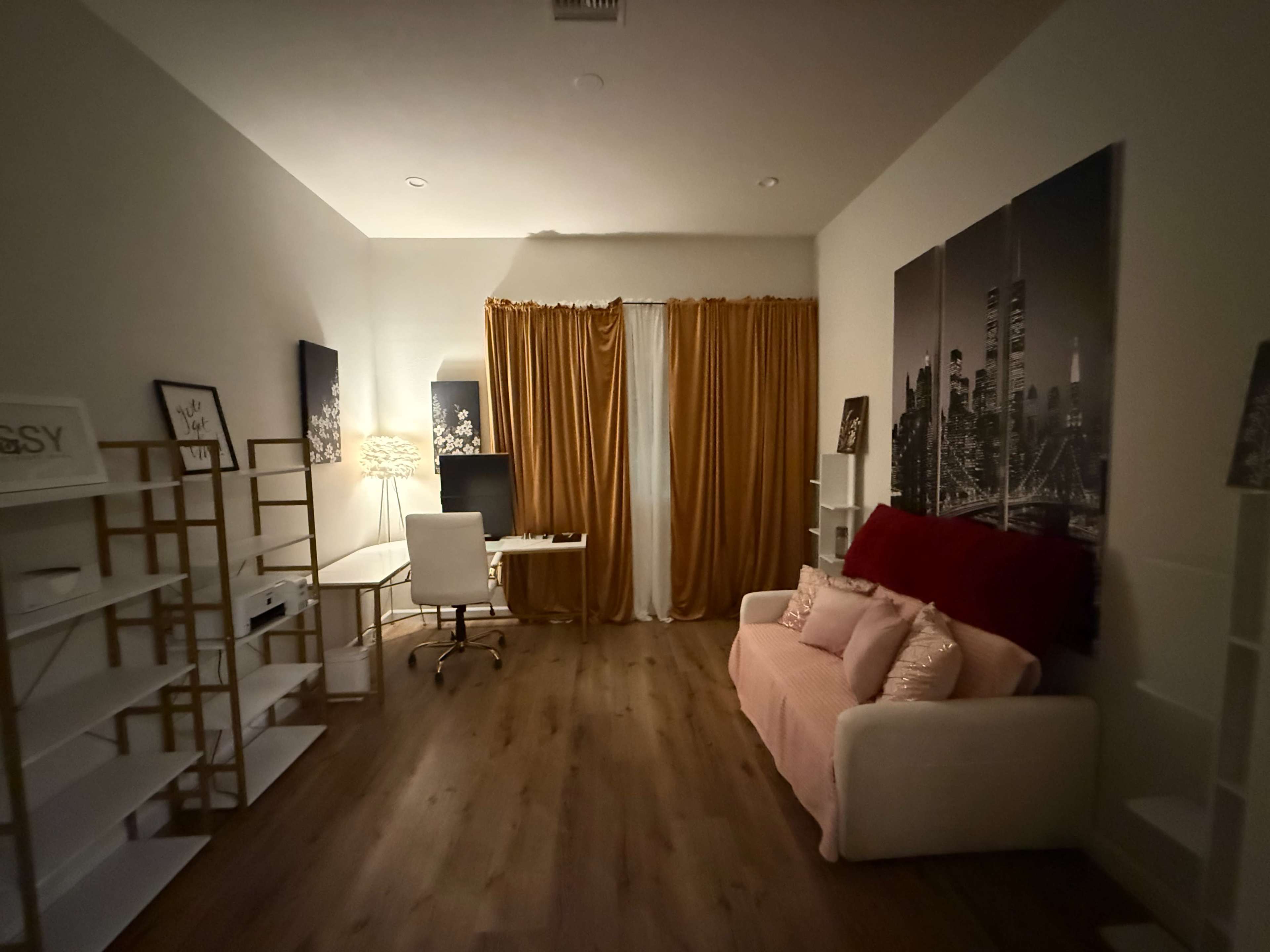The image shows a modern room featuring a pink sofa, a white desk with a computer, and tall curtains in a vibrant yellow hue, with shelving units on either side.