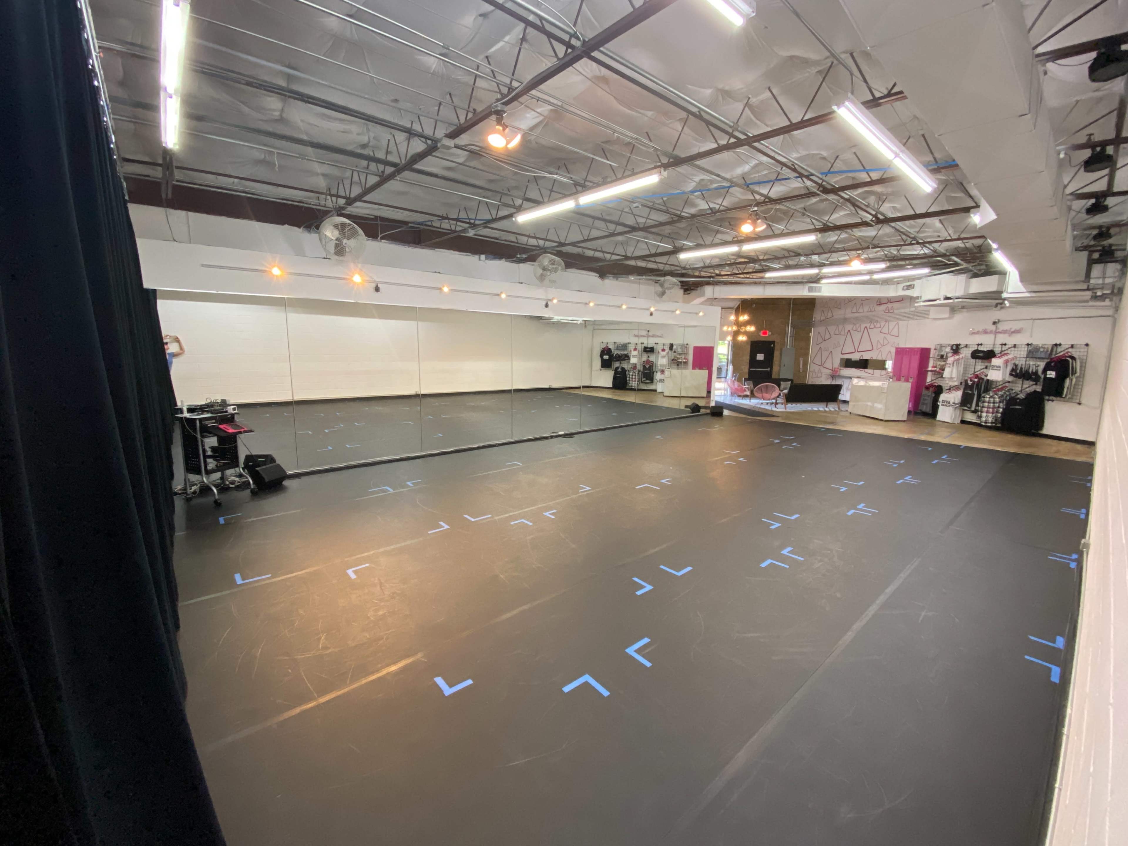 The image shows a spacious dance studio with a mirrored wall and a black floor marked with blue tape.