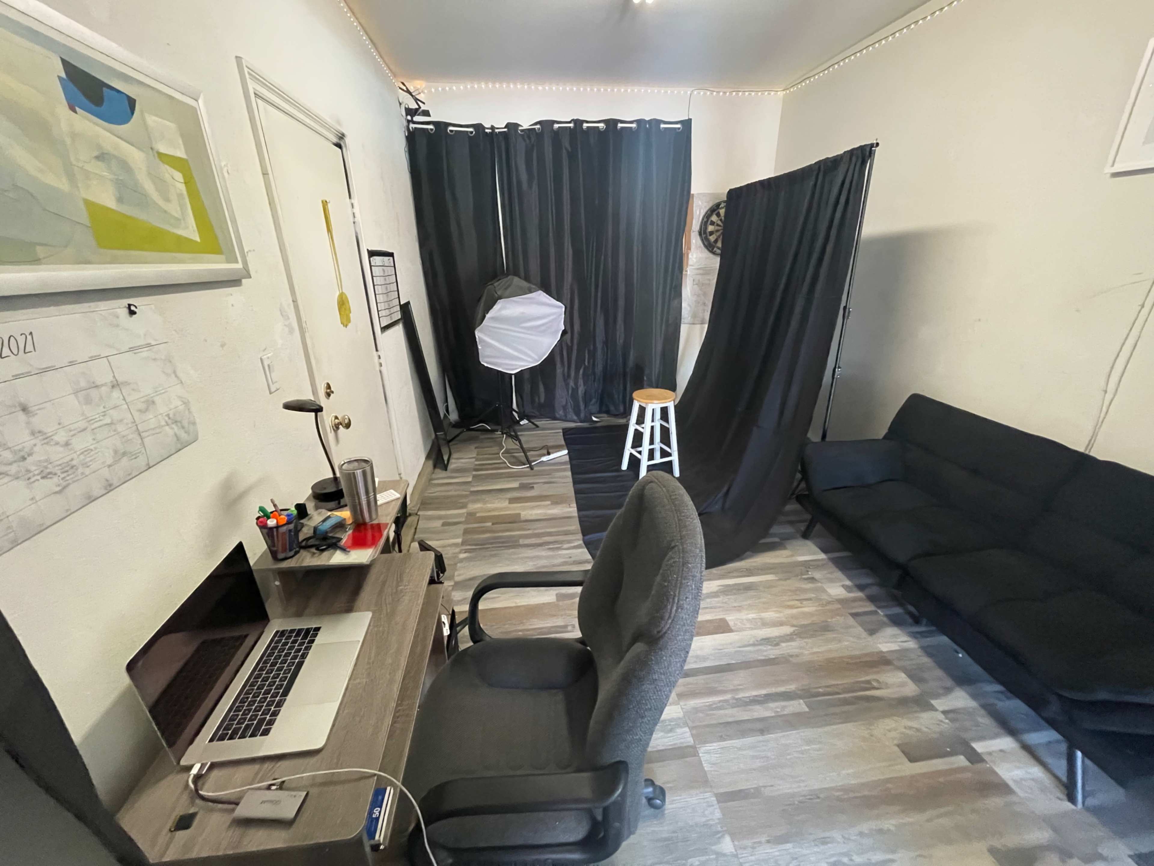 A small studio space with a desk and computer, a black backdrop and lighting equipment, and a white stool in the corner.