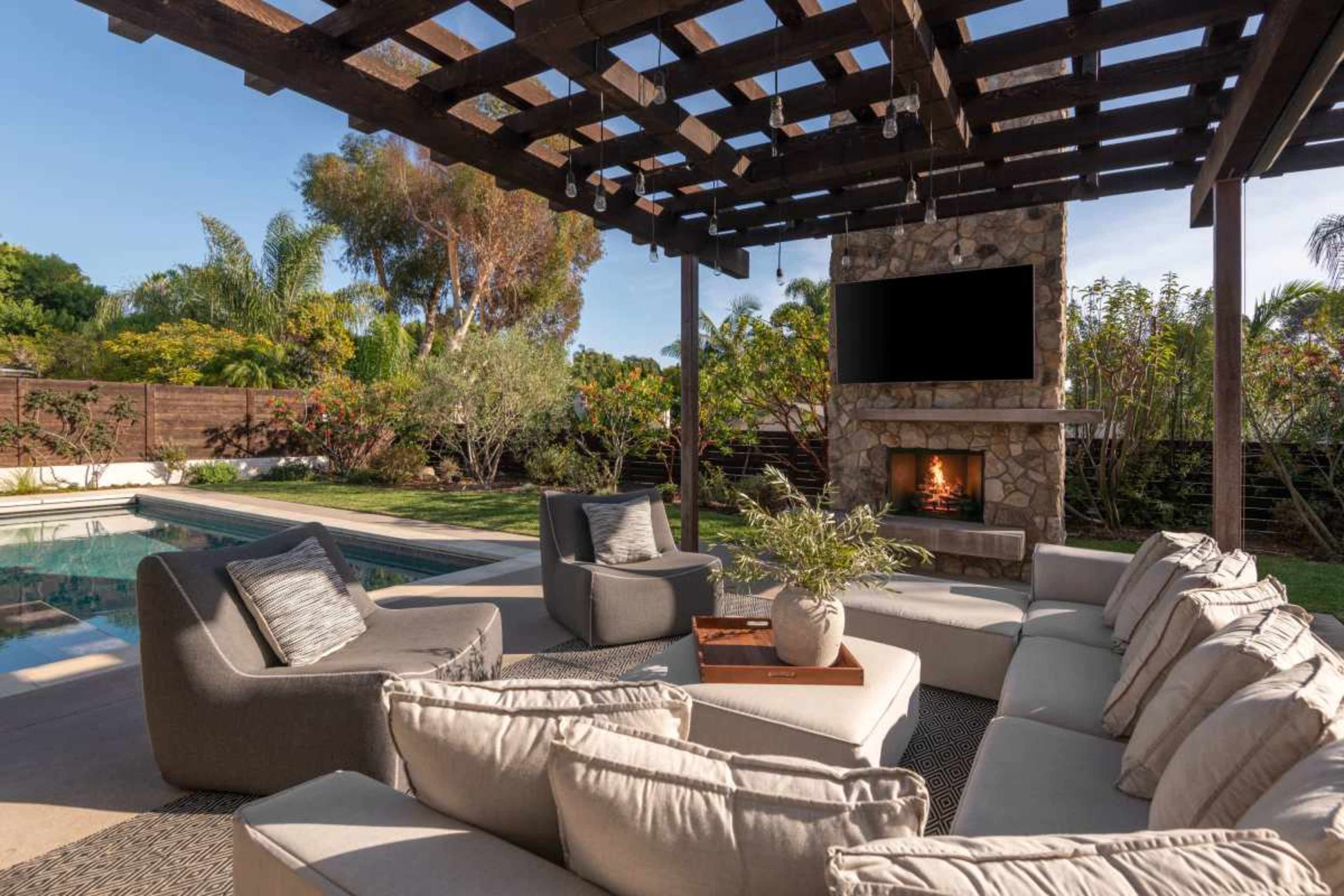 A comfortable outdoor seating area with a stone fireplace, television, and a pool surrounded by greenery.