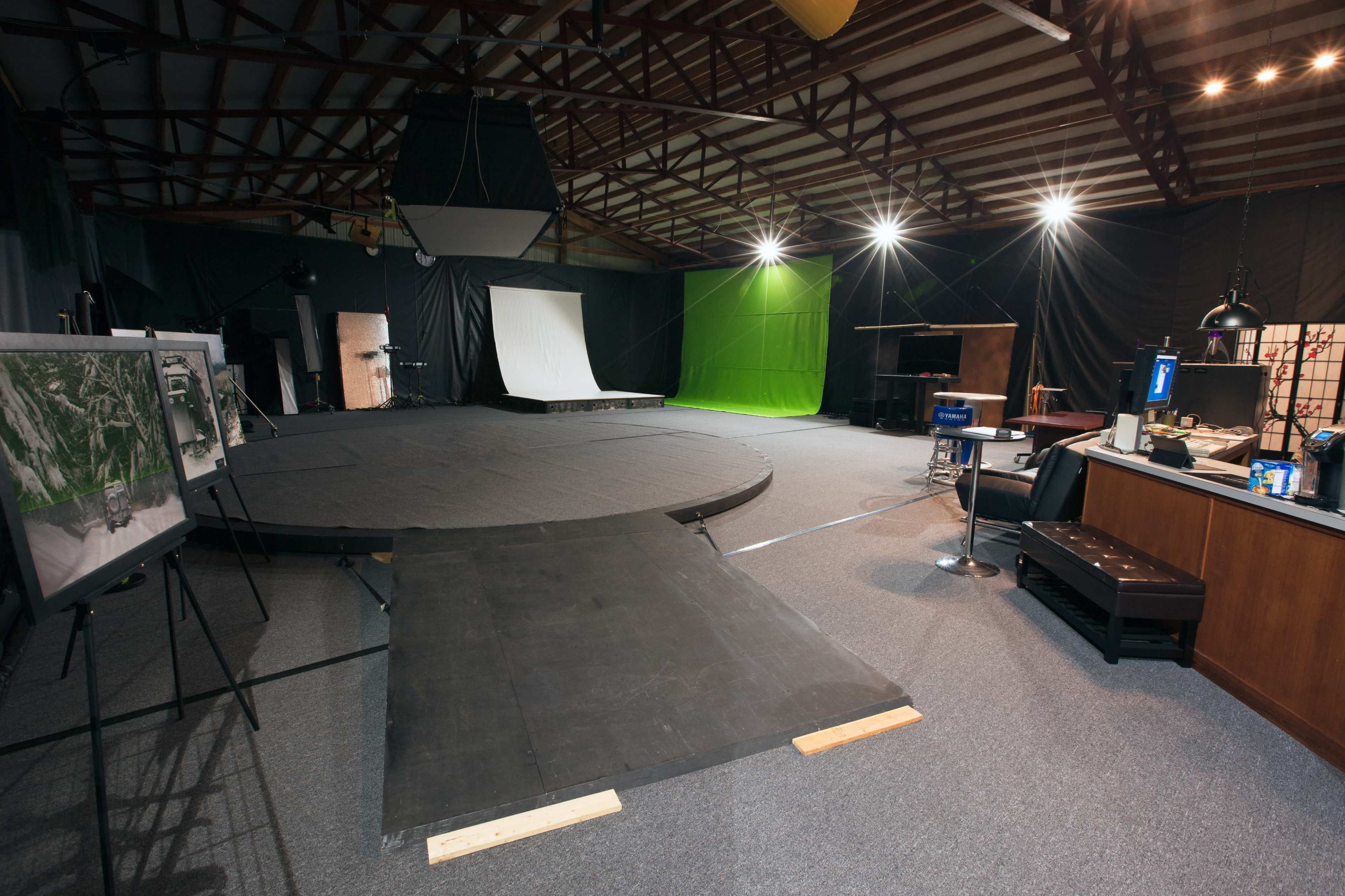 The image shows a spacious film studio setup with a circular stage, various backdrops, lighting equipment, and an observation area with furniture.