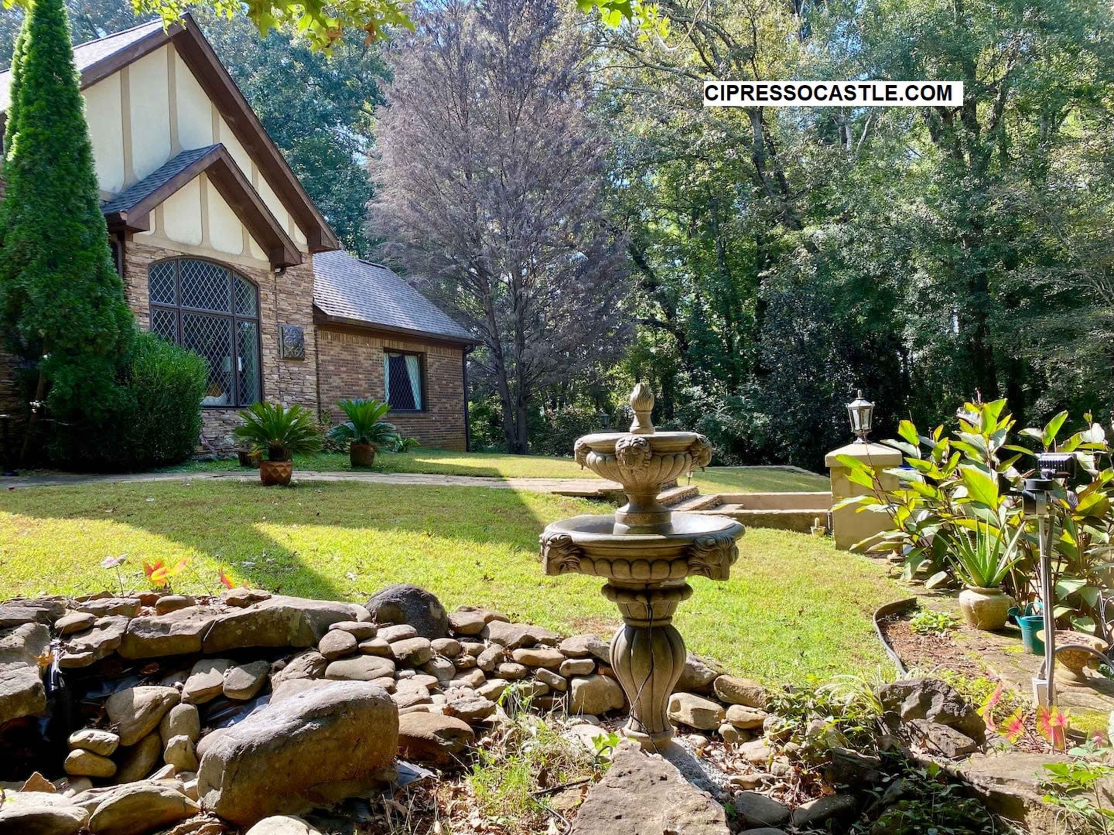 A stone fountain stands in the foreground of a landscaped yard, leading up to a house surrounded by trees.