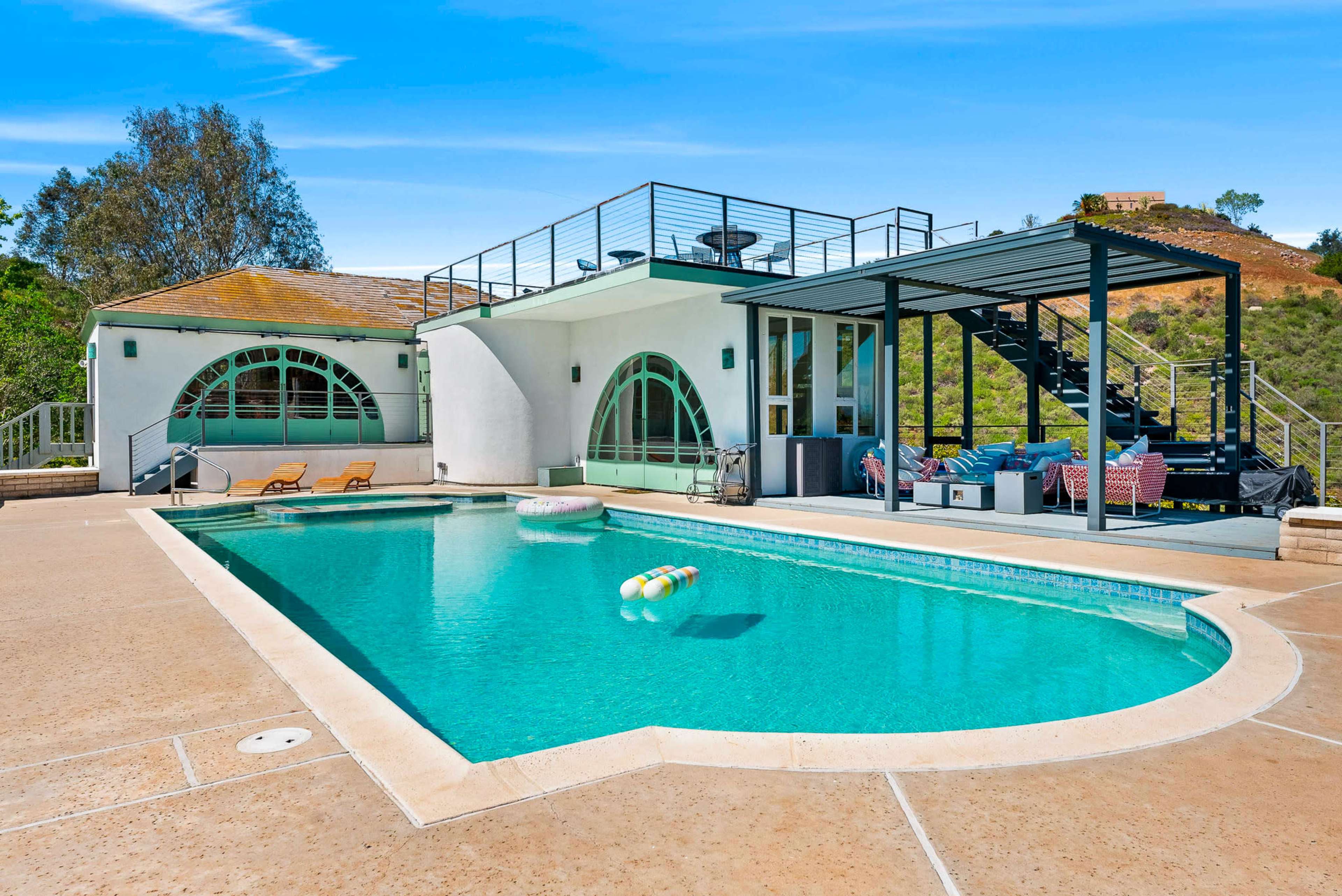 The image shows a modern swimming pool surrounded by a spacious patio area and a contemporary home with large arched windows.