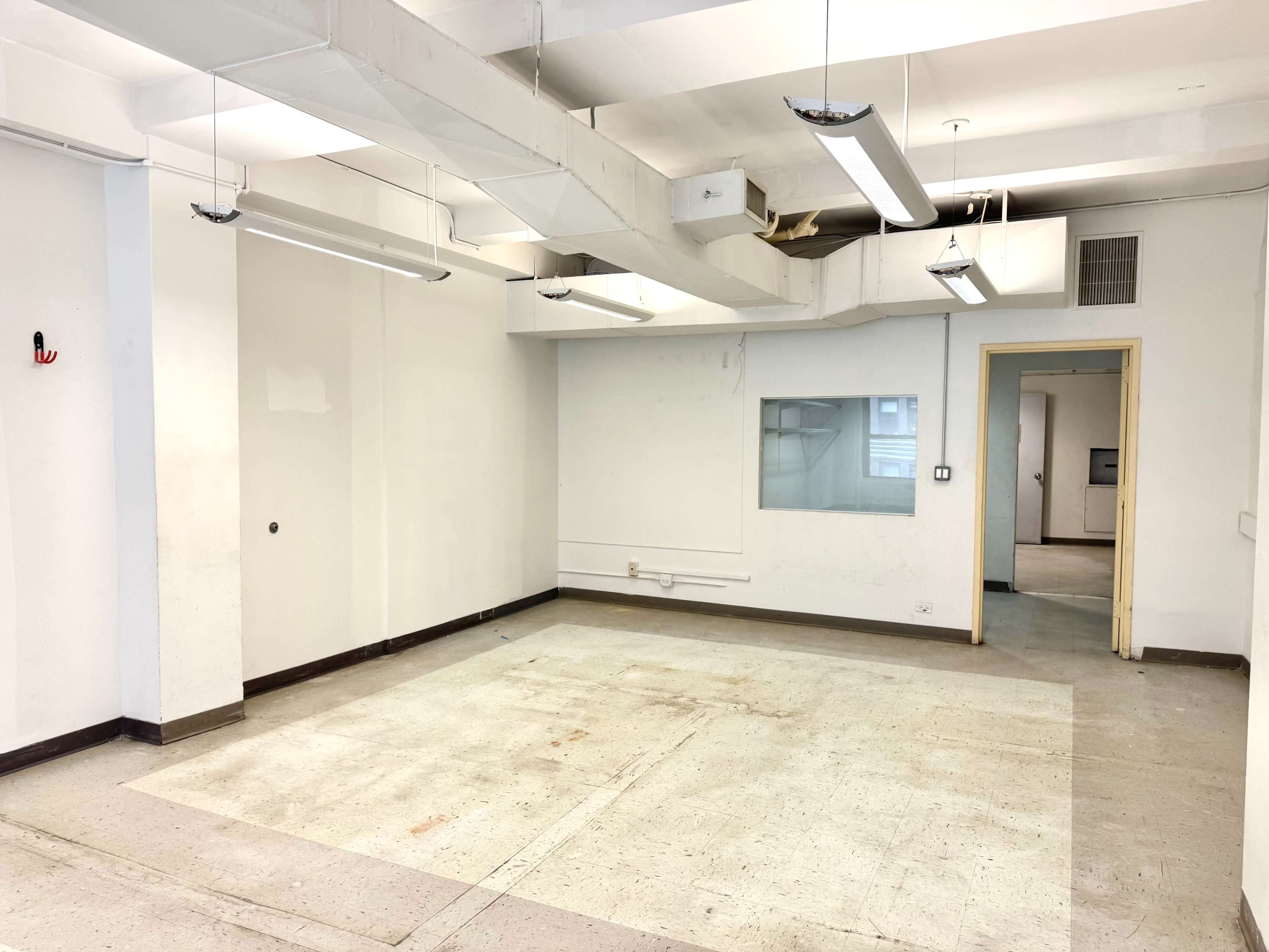 The image shows an empty room with concrete flooring, fluorescent lights, and exposed ductwork on the ceiling.