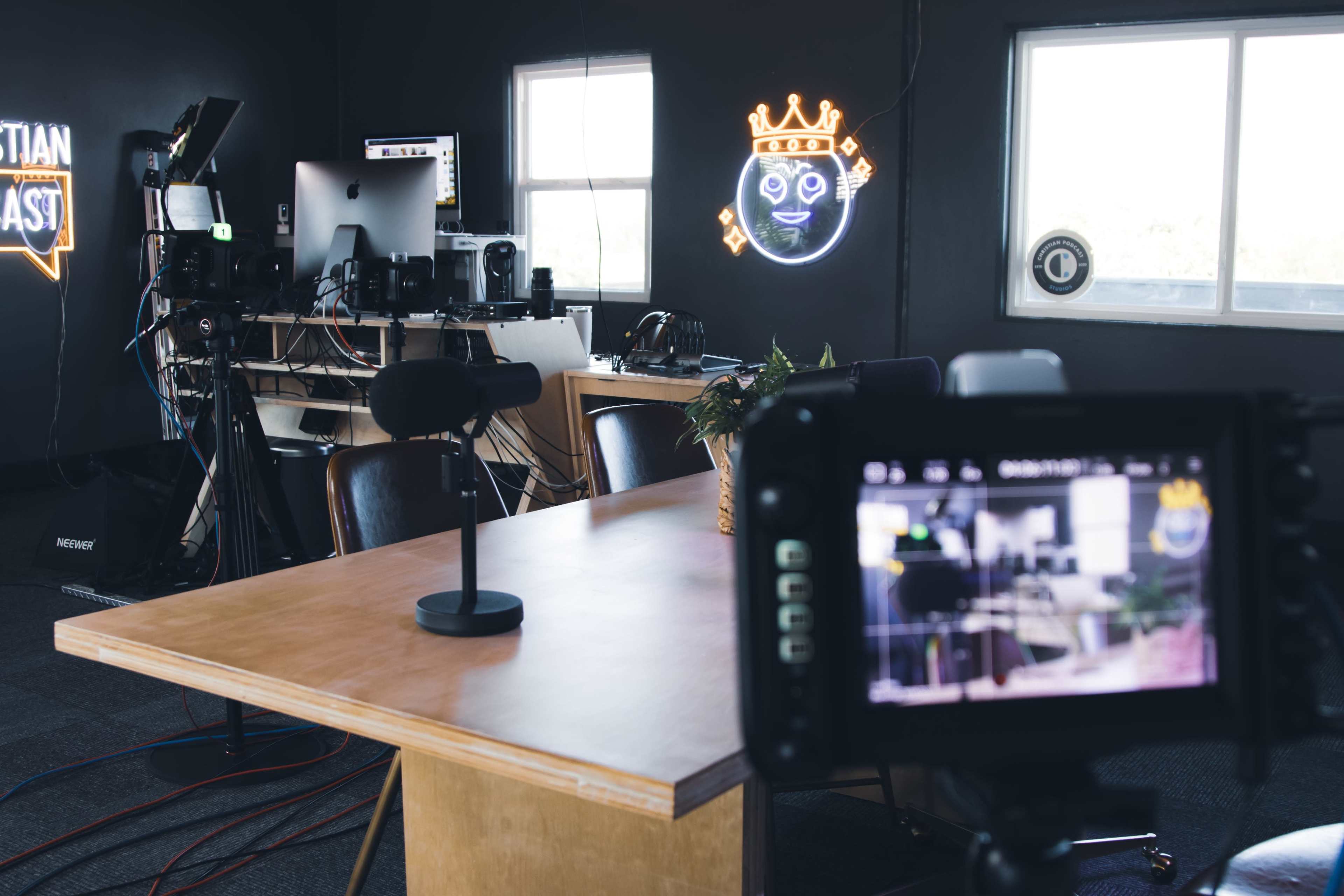 The image shows a modern podcast studio with a wooden table, a microphone, and various recording equipment, along with a neon sign on the wall.