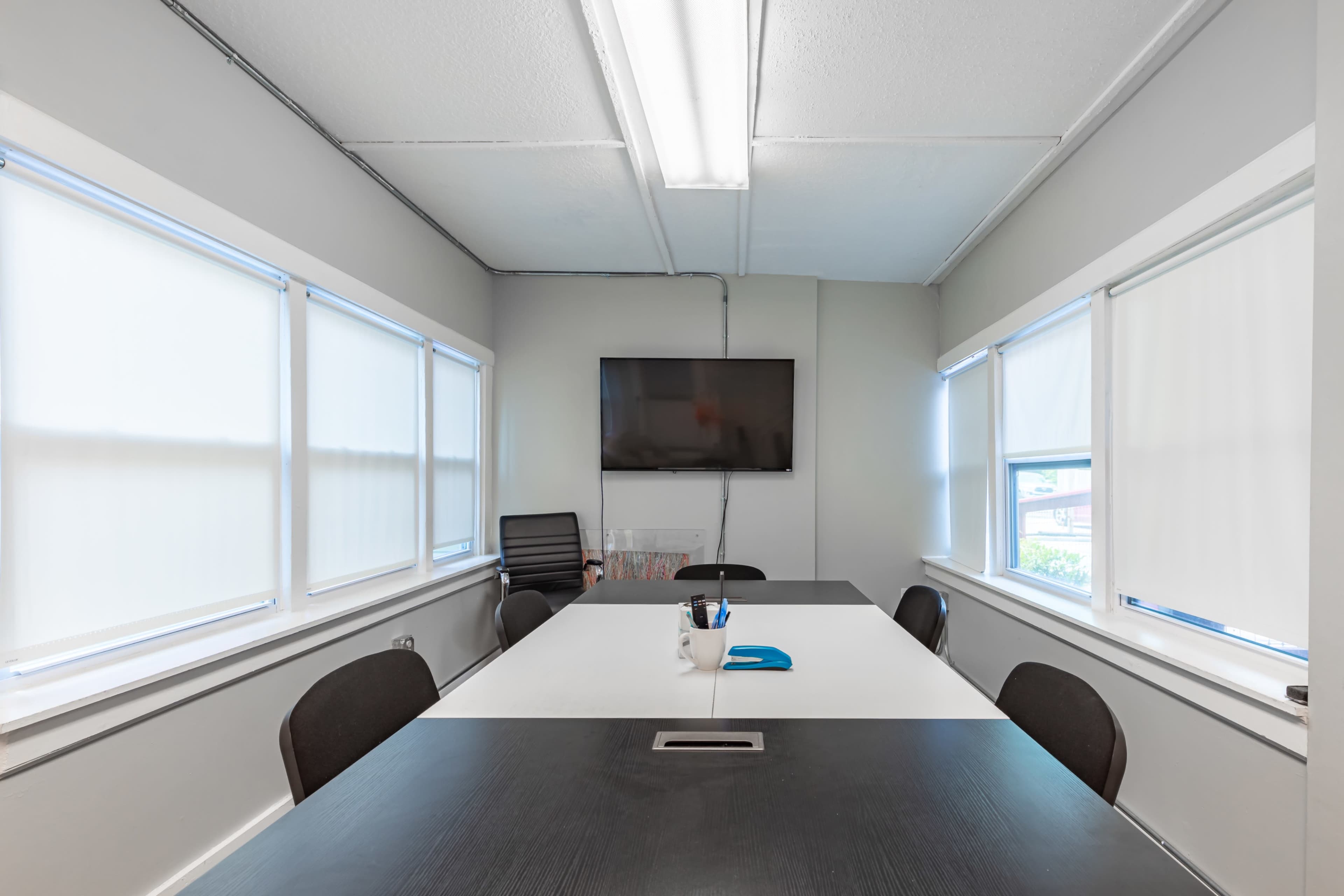 A conference room features a large table, four chairs, a wall-mounted TV, and windows with blinds.
