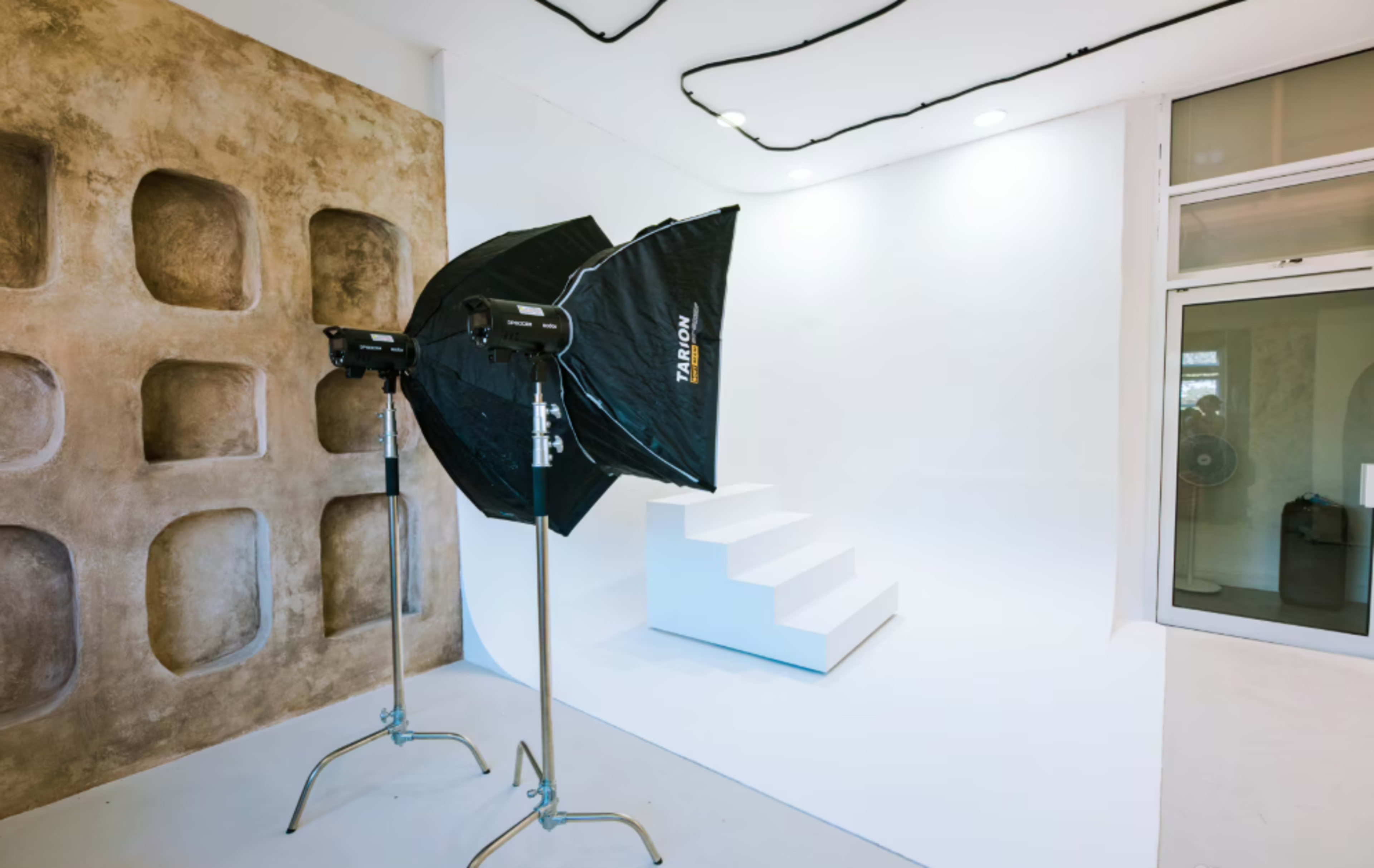The image shows a photography studio featuring a white background, stairs, and lighting equipment set up for photo shoots.