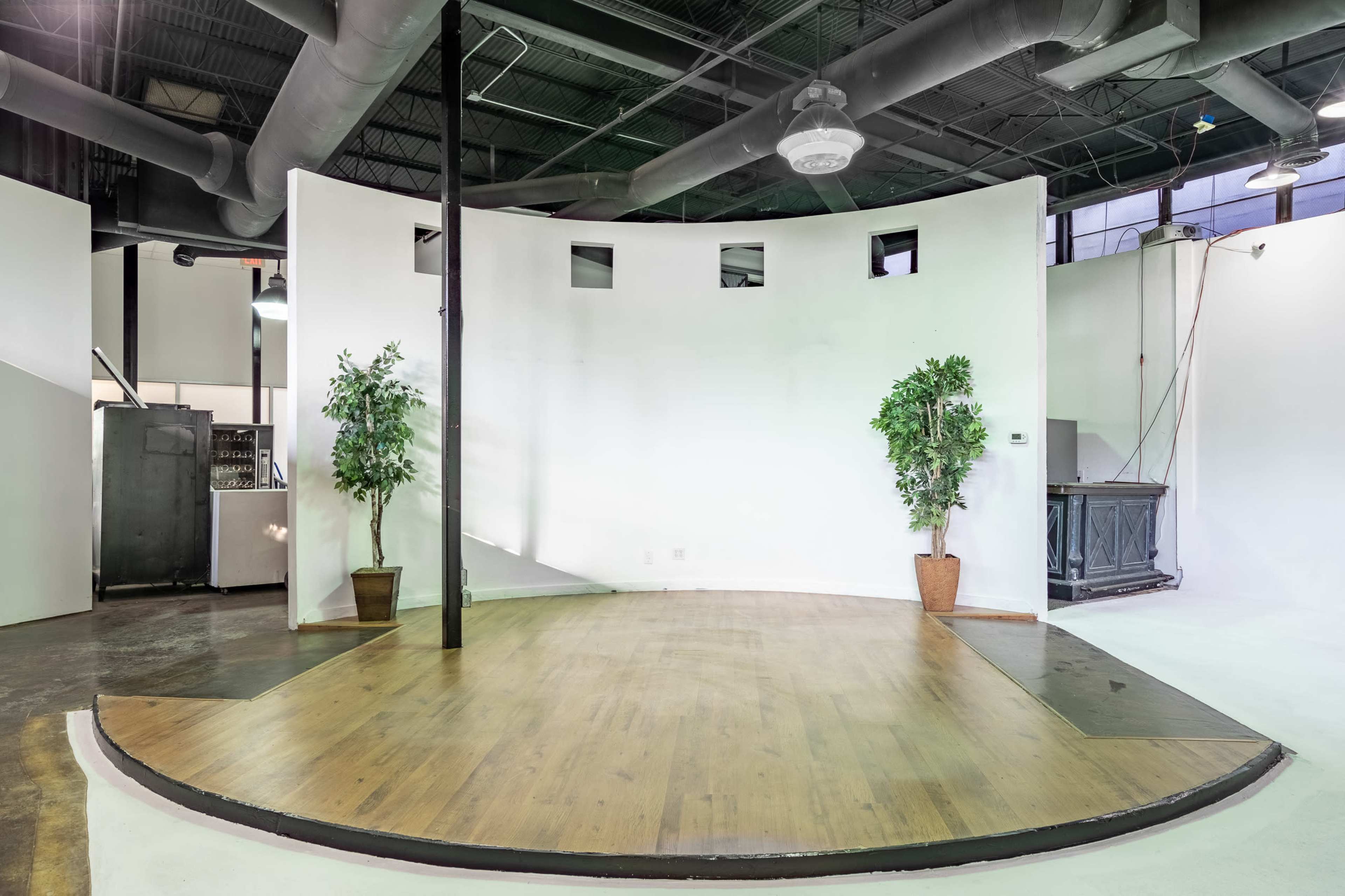 The image shows a bare stage with hardwood flooring, two potted plants on either side, and a white backdrop in an industrial space.