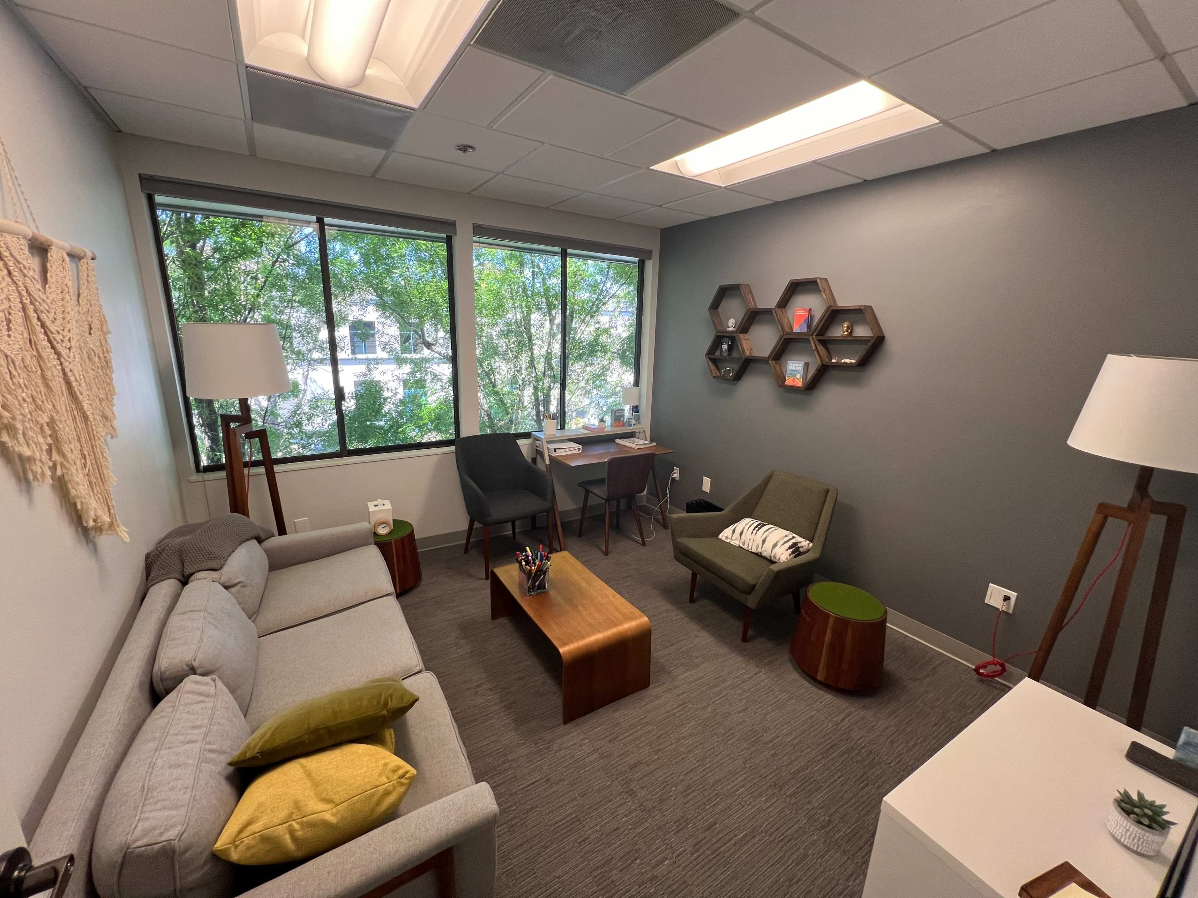 A cozy office space features a grey sofa, two chairs, a wooden coffee table, and a desk, with large windows allowing natural light and greenery to enter.