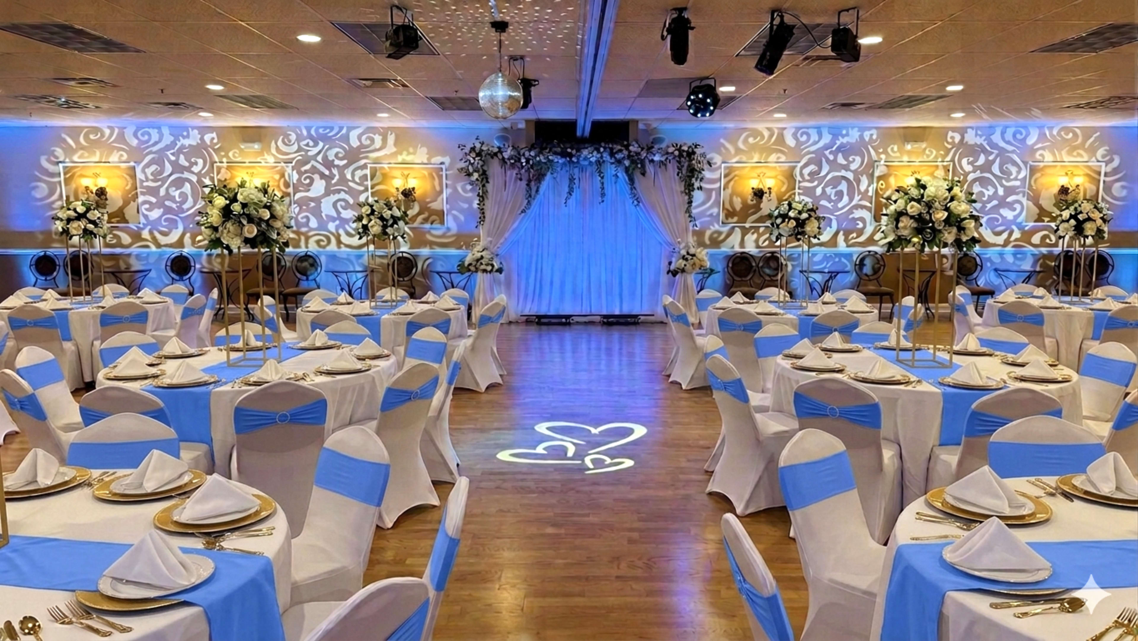 The image shows a banquet hall set for an event, with tables covered in white cloths and blue accents, arranged around a decorated stage featuring floral arrangements and draped fabric.