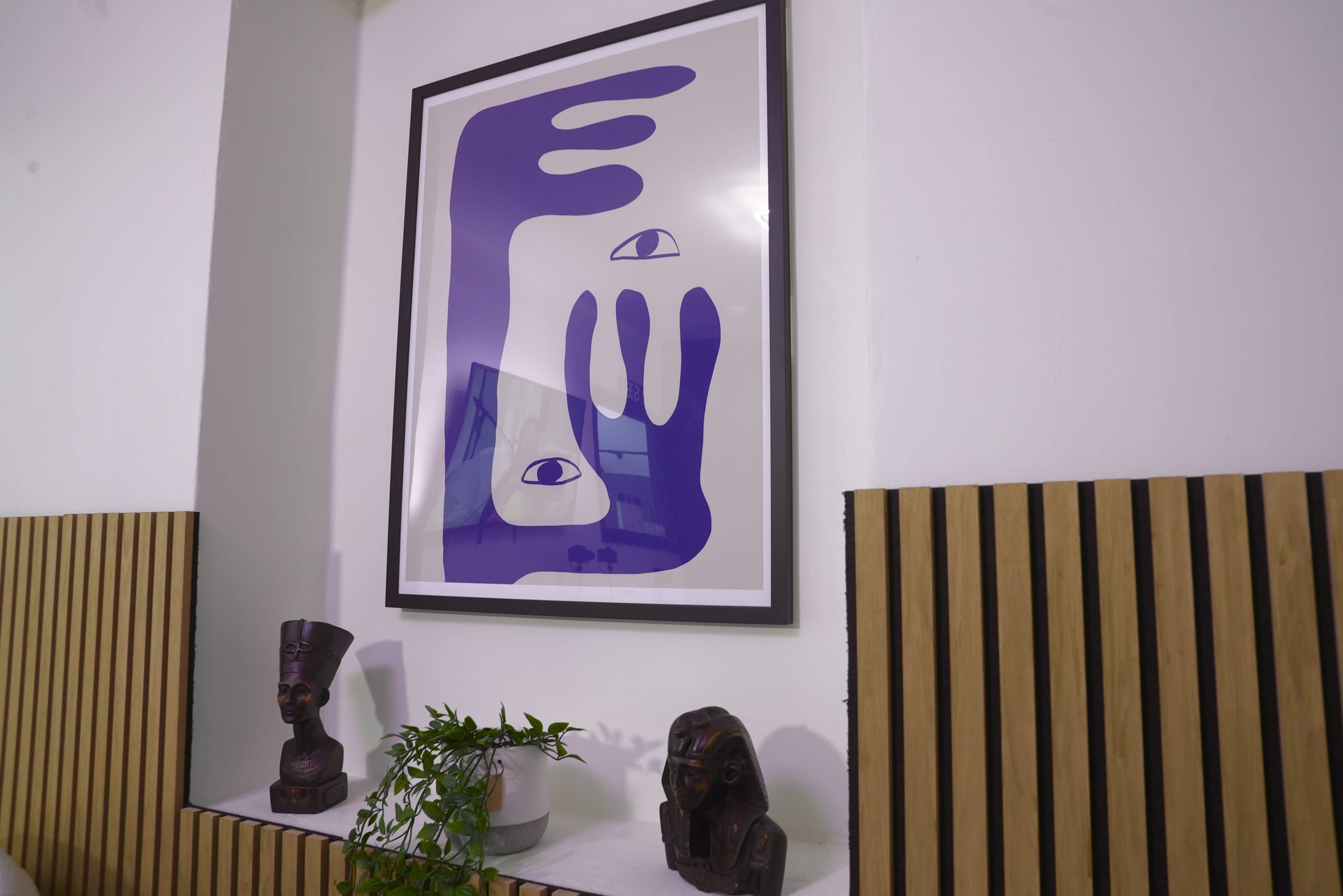 A framed abstract artwork featuring purple hands and eyes is displayed on a white wall above a wooden shelf with decorative sculptures and a small plant.