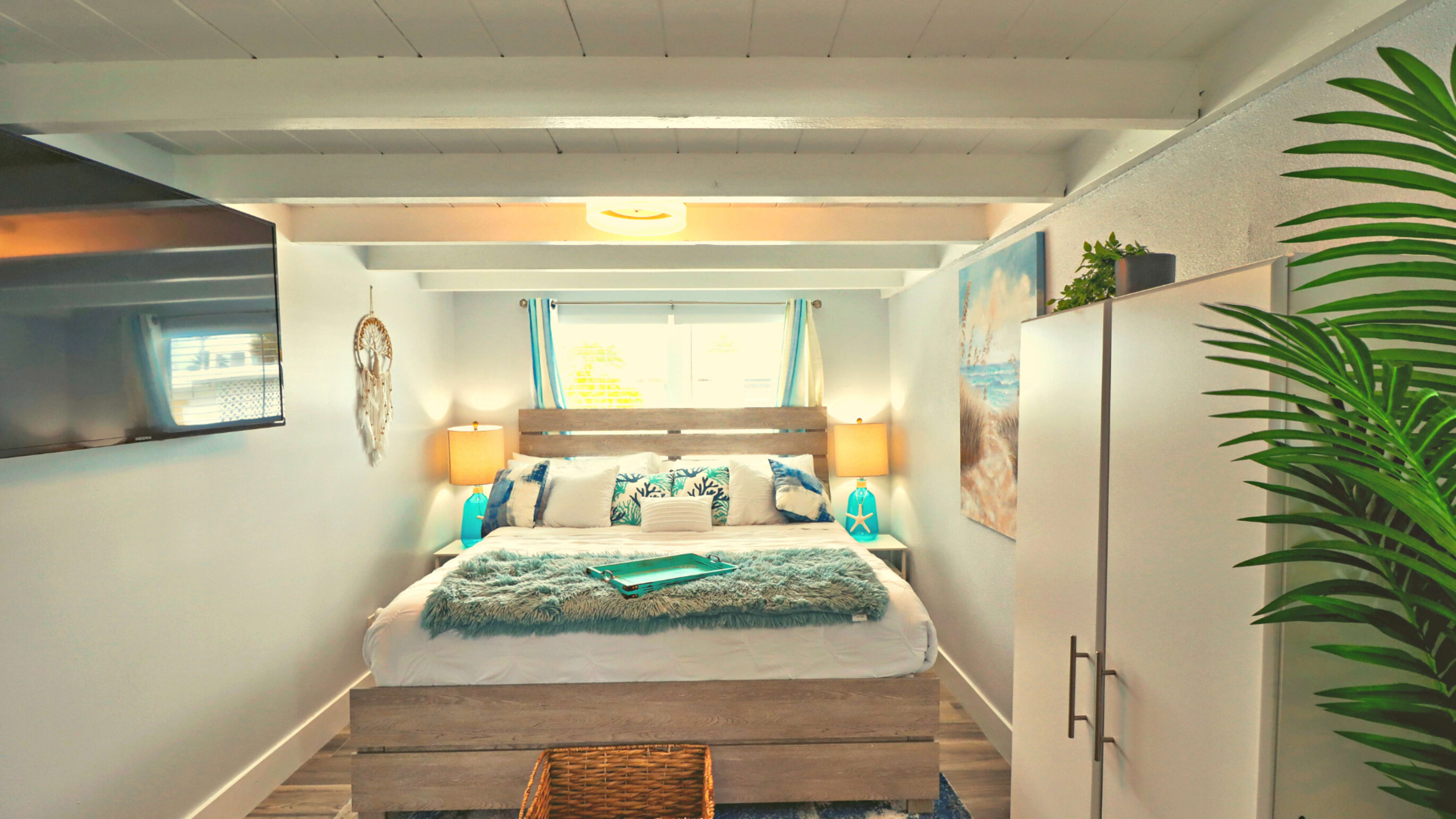 The bedroom features a bed with a wooden headboard, flanked by two lamps, a television mounted on the wall, and a large window dressed with blue curtains.