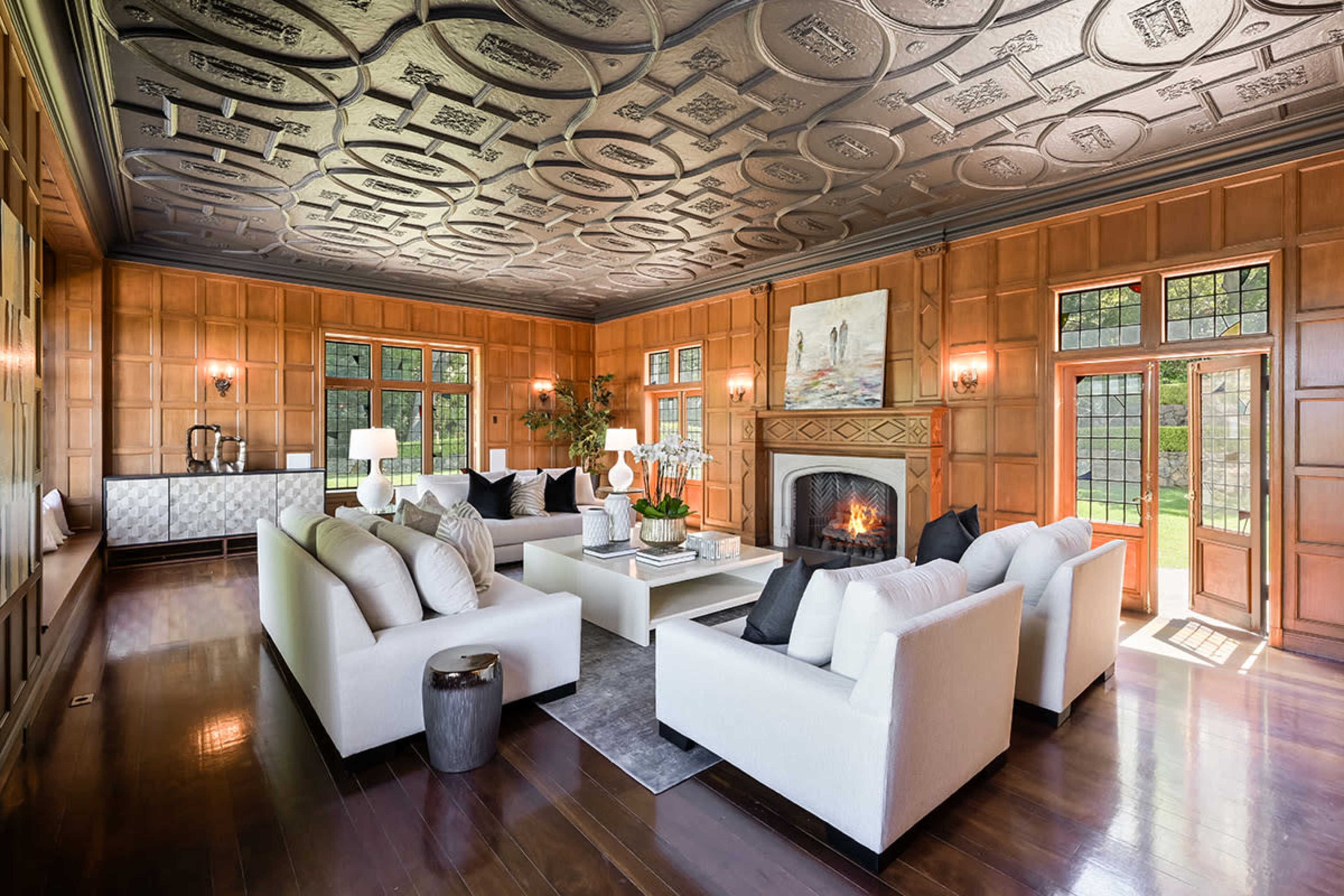 The image shows a spacious living room with a high, decorative ceiling, large windows, and two white sofas arranged around a central coffee table, featuring a fireplace and wooden paneling on the walls.