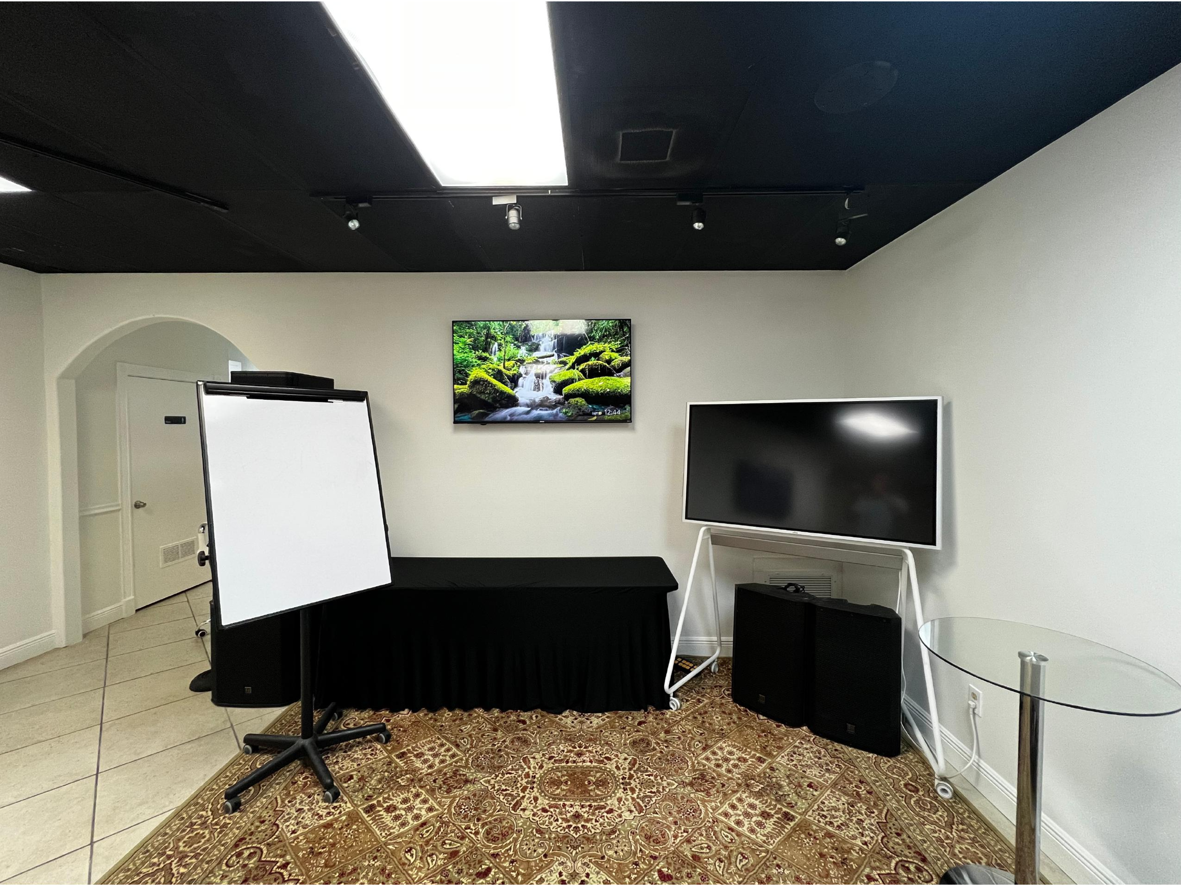 The image shows a meeting room with a large screen displaying a nature scene, a presentation whiteboard on a stand, and a glass table beside two audio speakers.