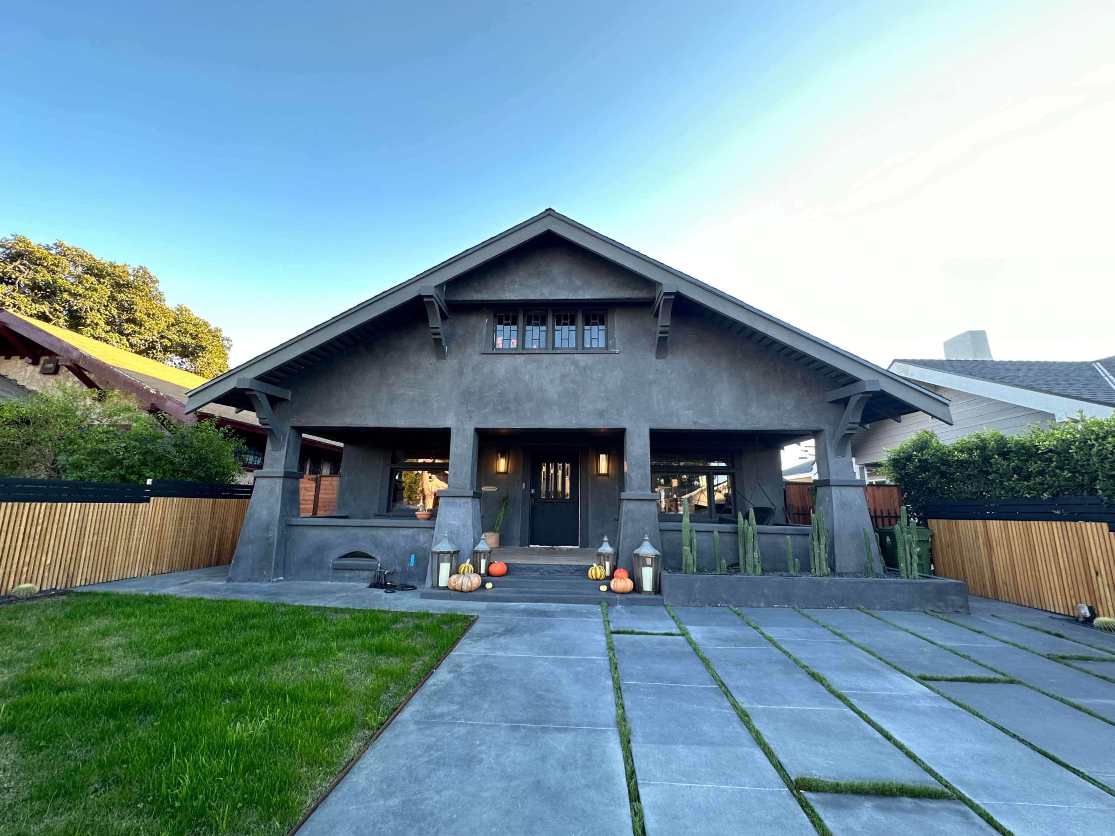 A modern, gray stucco house with a steep roof and a stone pathway is set in a landscaped yard featuring pumpkins and decorative elements.