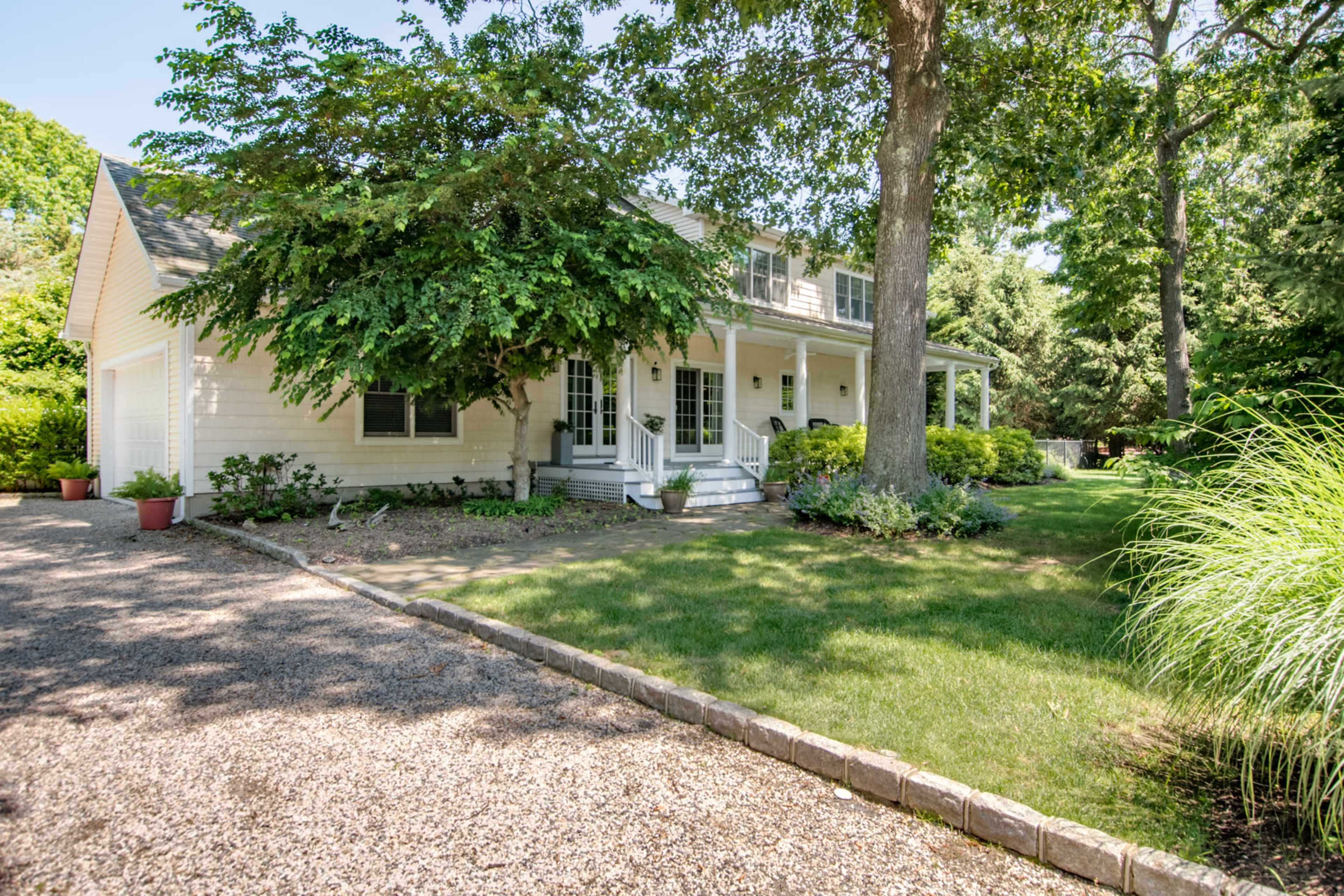 A two-story house with a covered porch and a gravel driveway is surrounded by green trees and landscaped shrubs.