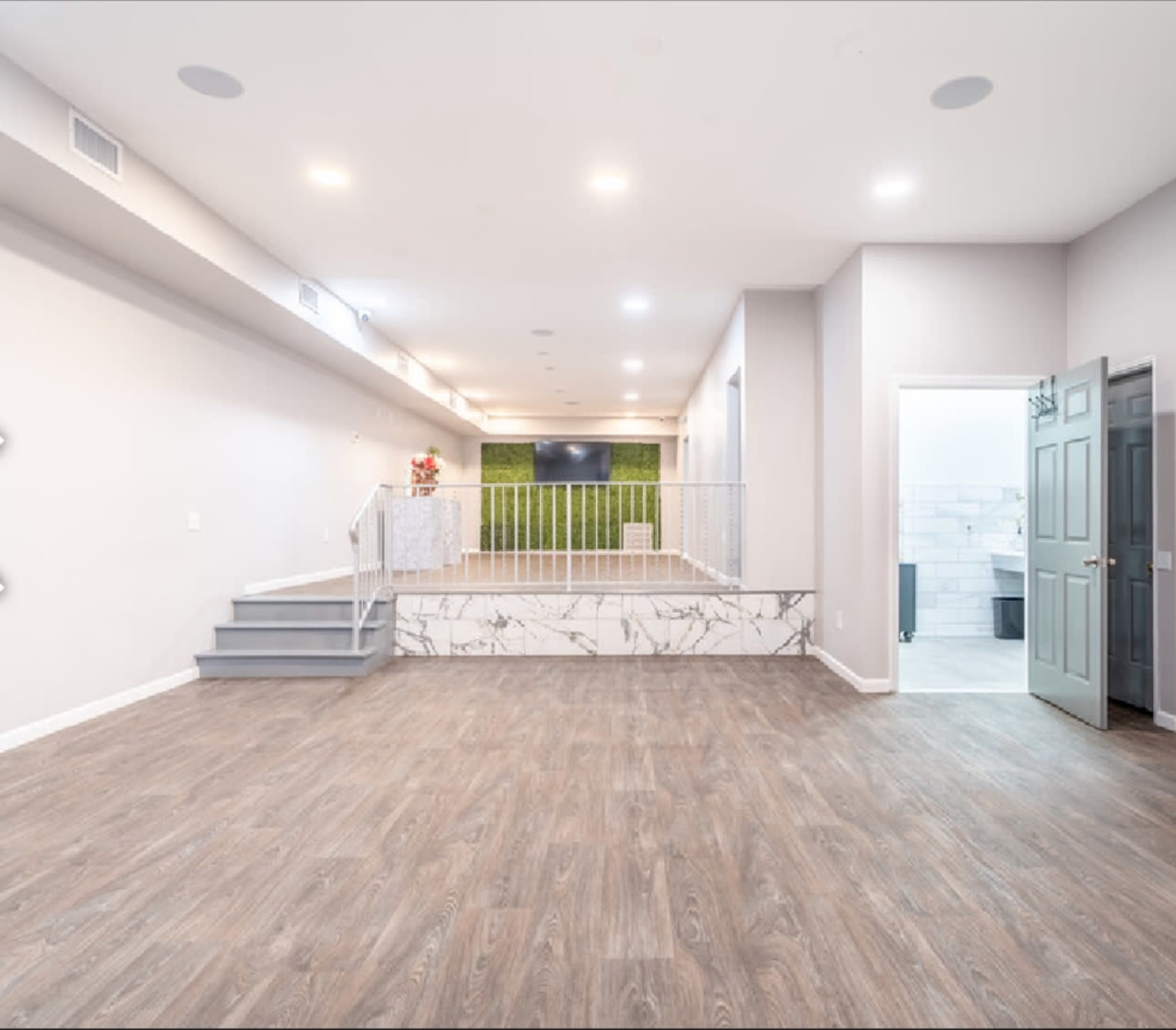 A spacious, well-lit room with wooden flooring, featuring a staircase, a green wall, and a doorway leading to a bathroom area.