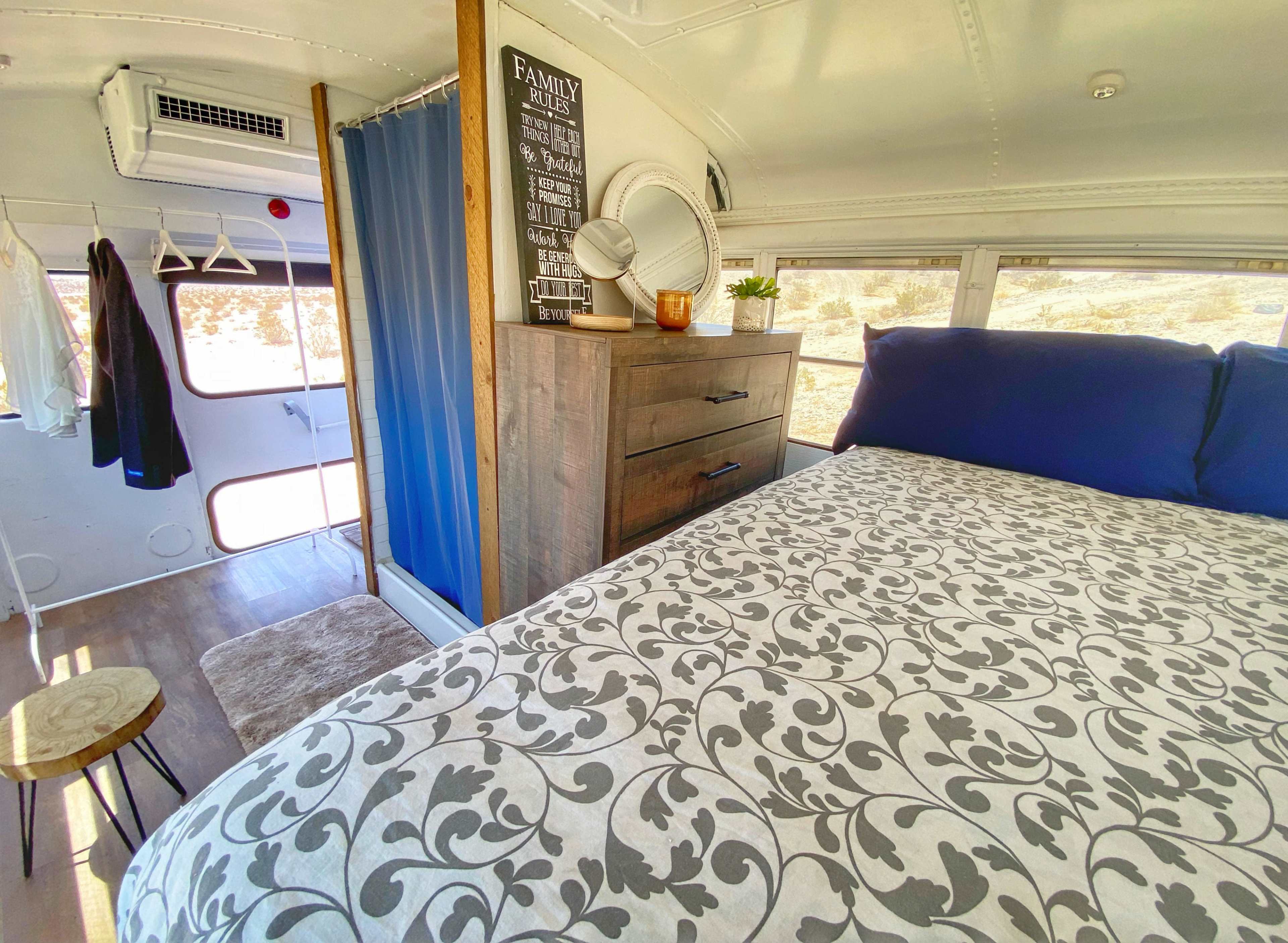 The image shows the interior of a converted bus featuring a bed with a patterned duvet, a dresser, a mirror, and a curtain partitioning the space.