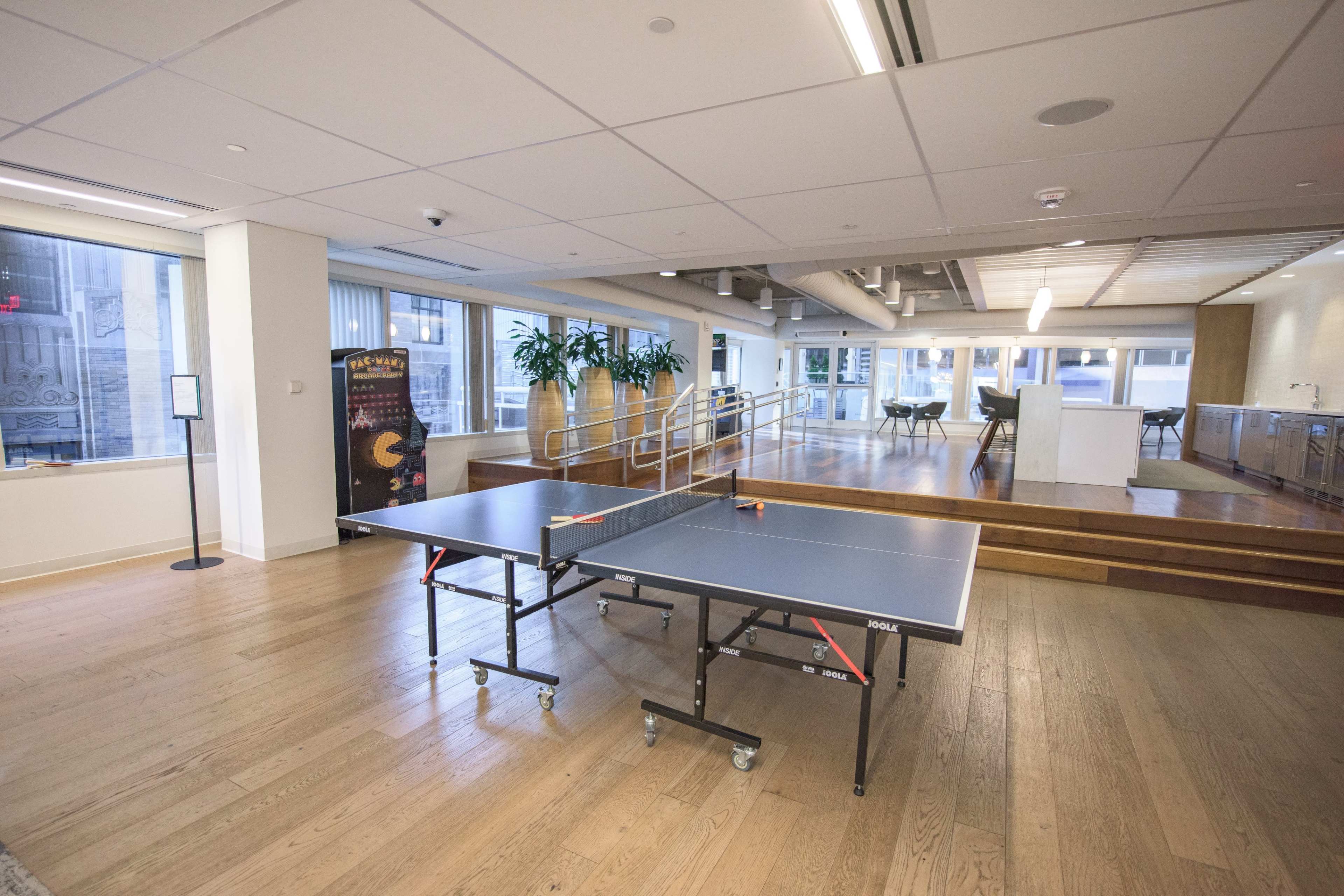 A ping pong table is centrally positioned in a spacious room with large windows and wooden flooring.