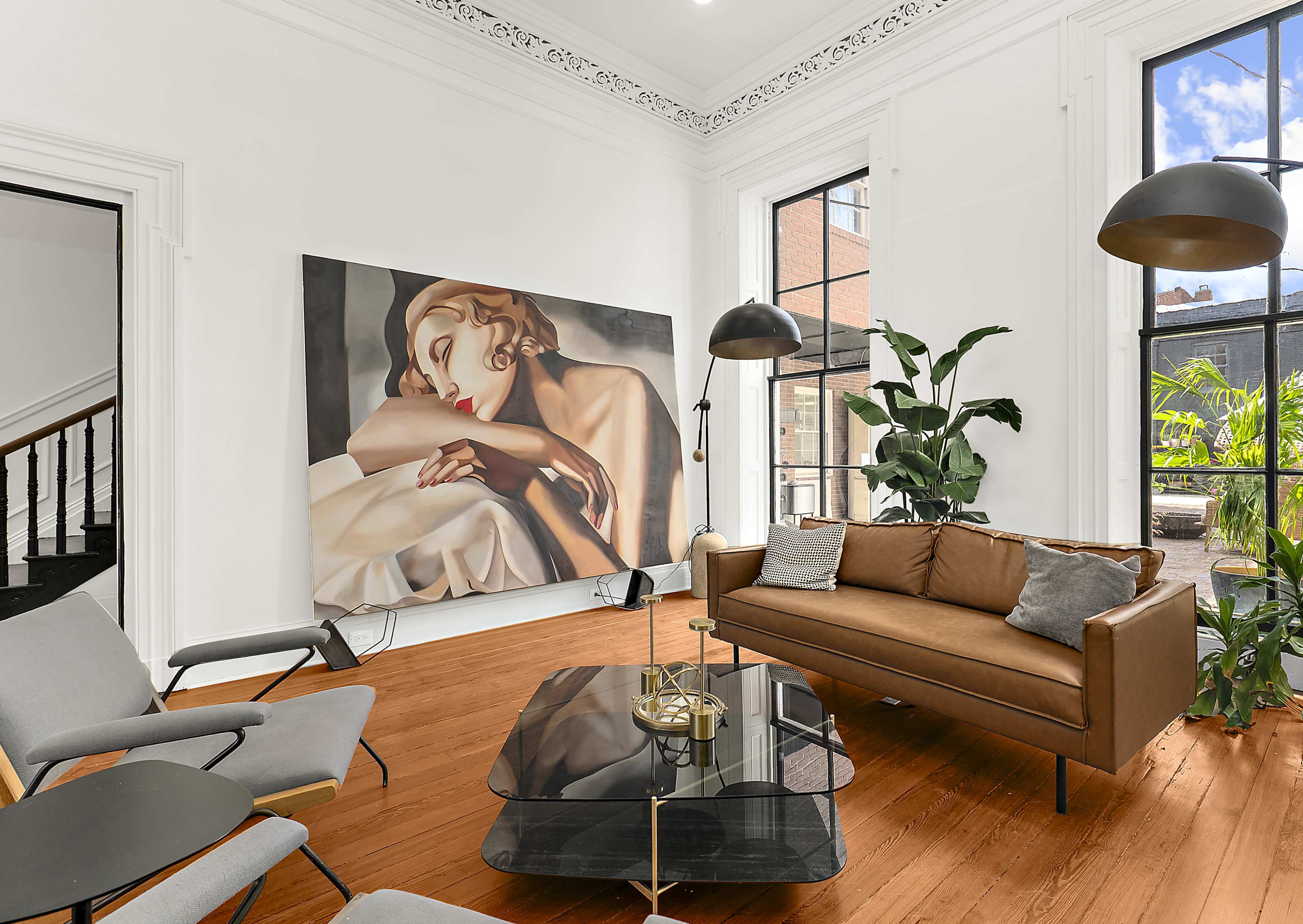 The image shows a modern living room with a brown sofa, a glass coffee table, two armchairs, a large painting on the wall, and windows that let in natural light, surrounded by indoor plants.