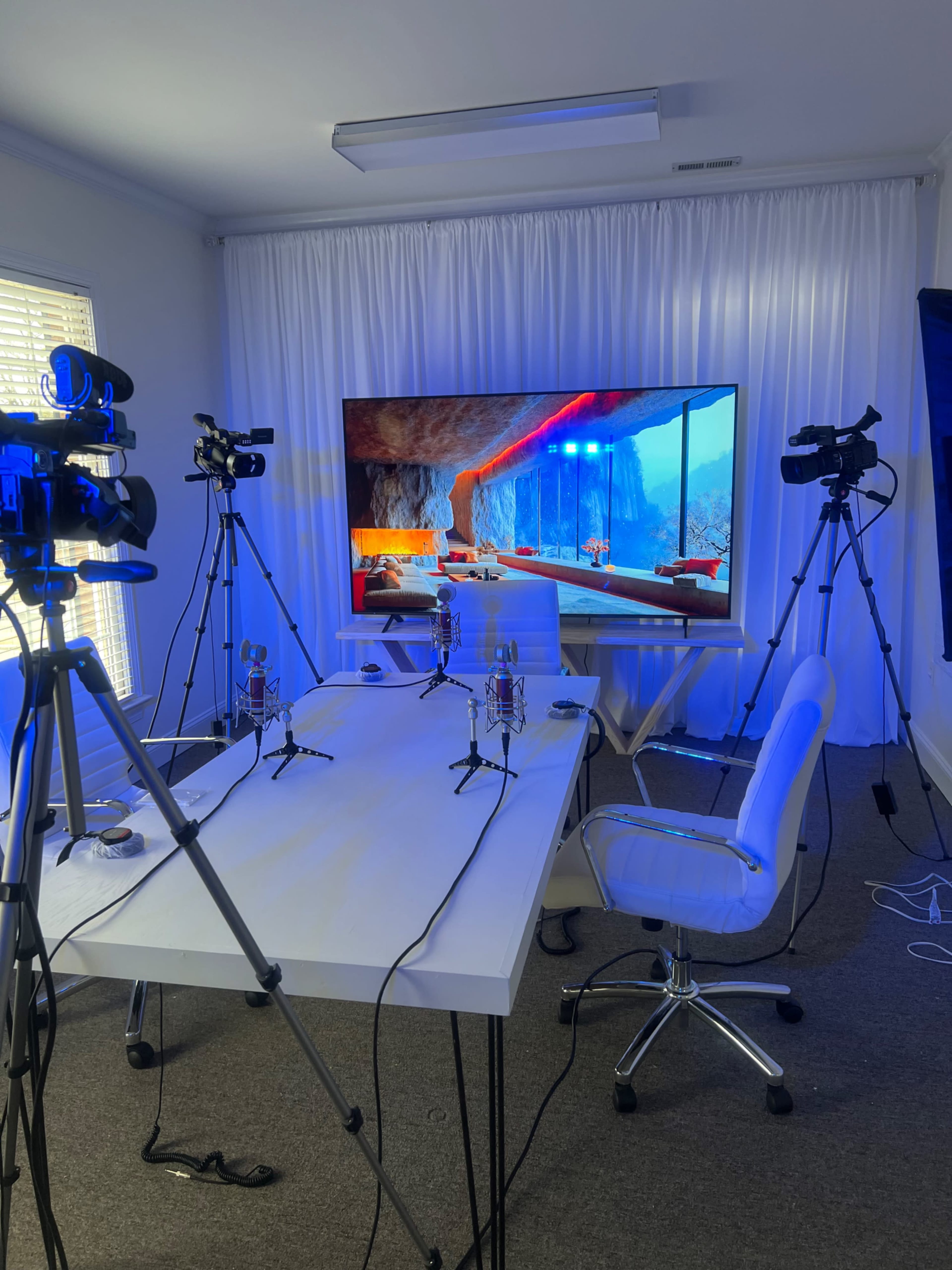 A modern video recording setup featuring a white table with microphones, two cameras on tripods, and a large screen displaying a colorful scene, all set in a well-lit room with white curtains.