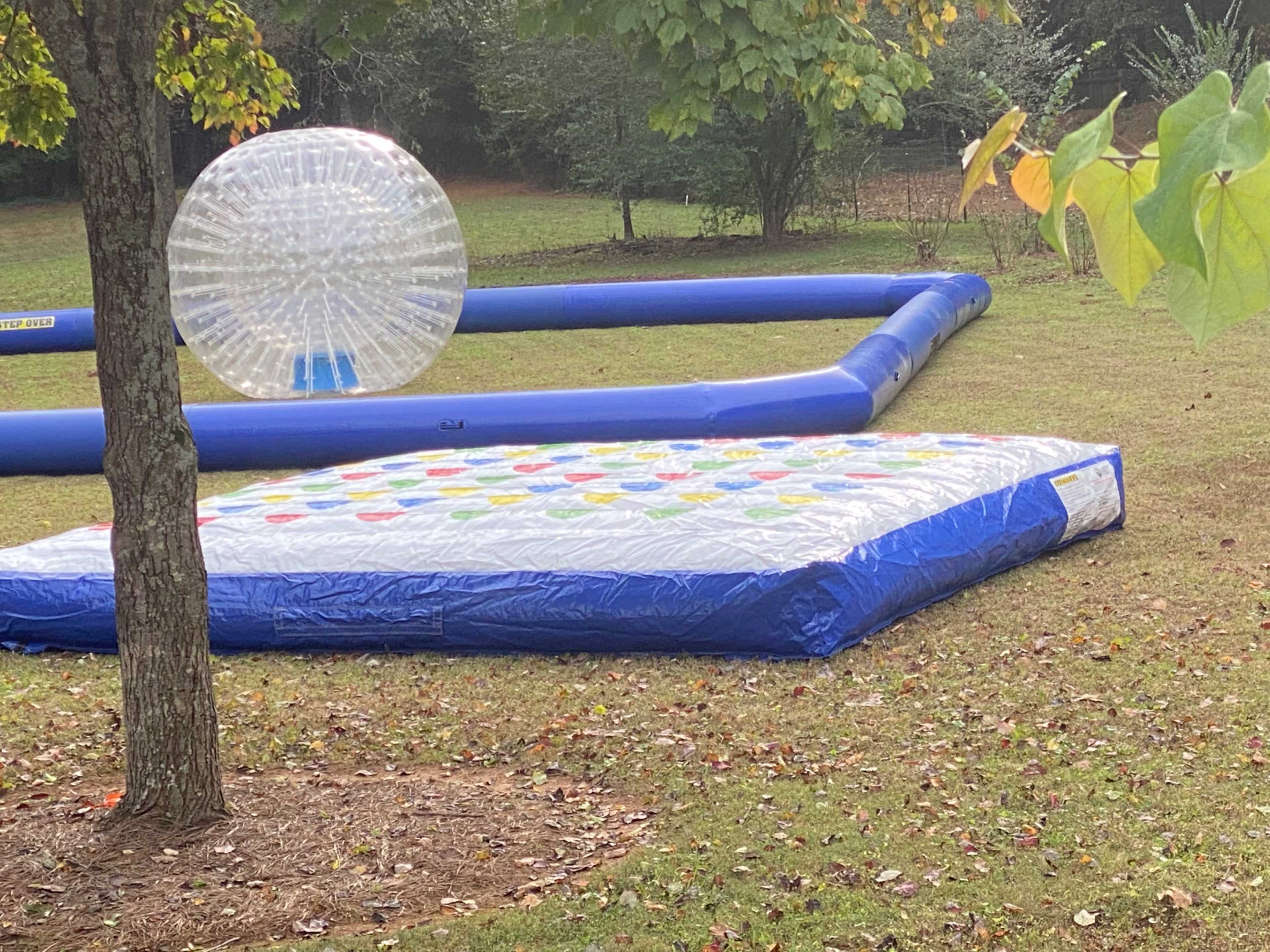 The image shows an inflatable water attraction featuring a colorful, circular pad and a large, transparent inflatable sphere placed on a grassy area surrounded by trees.