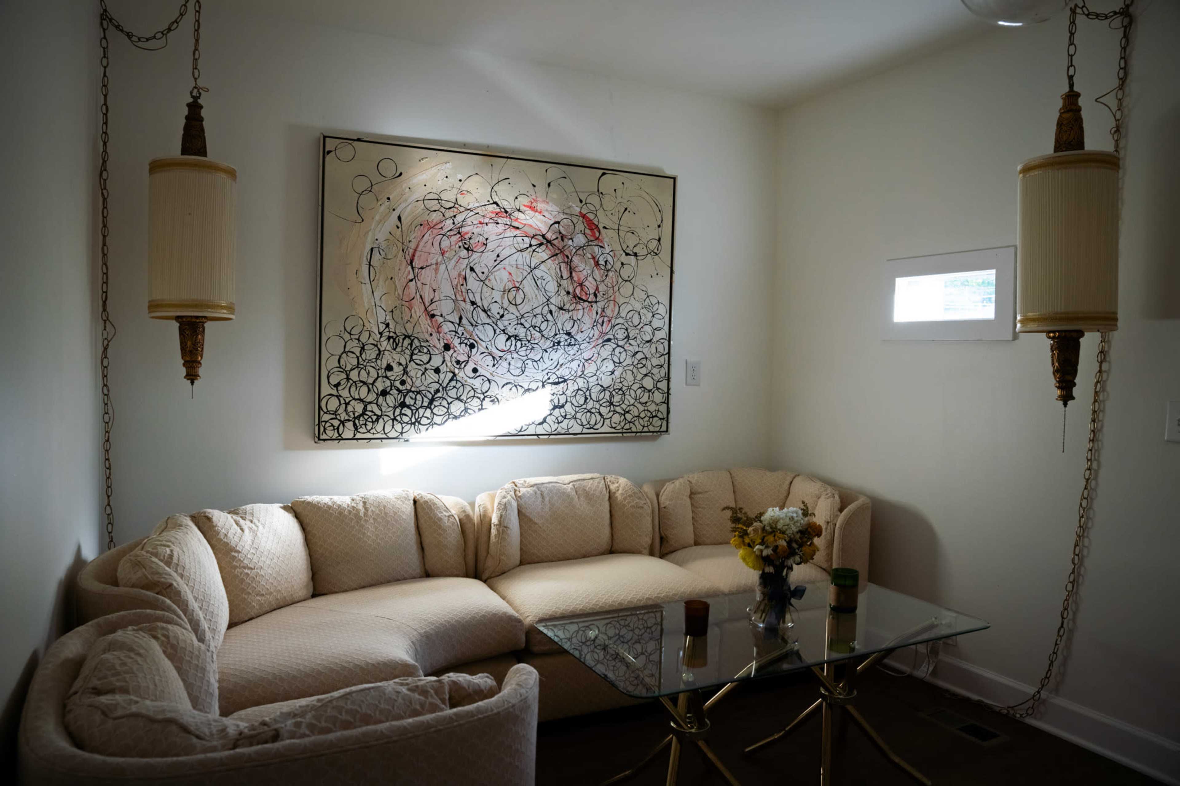 A curved beige sofa is positioned around a glass coffee table in a well-lit room featuring an abstract painting on the wall.