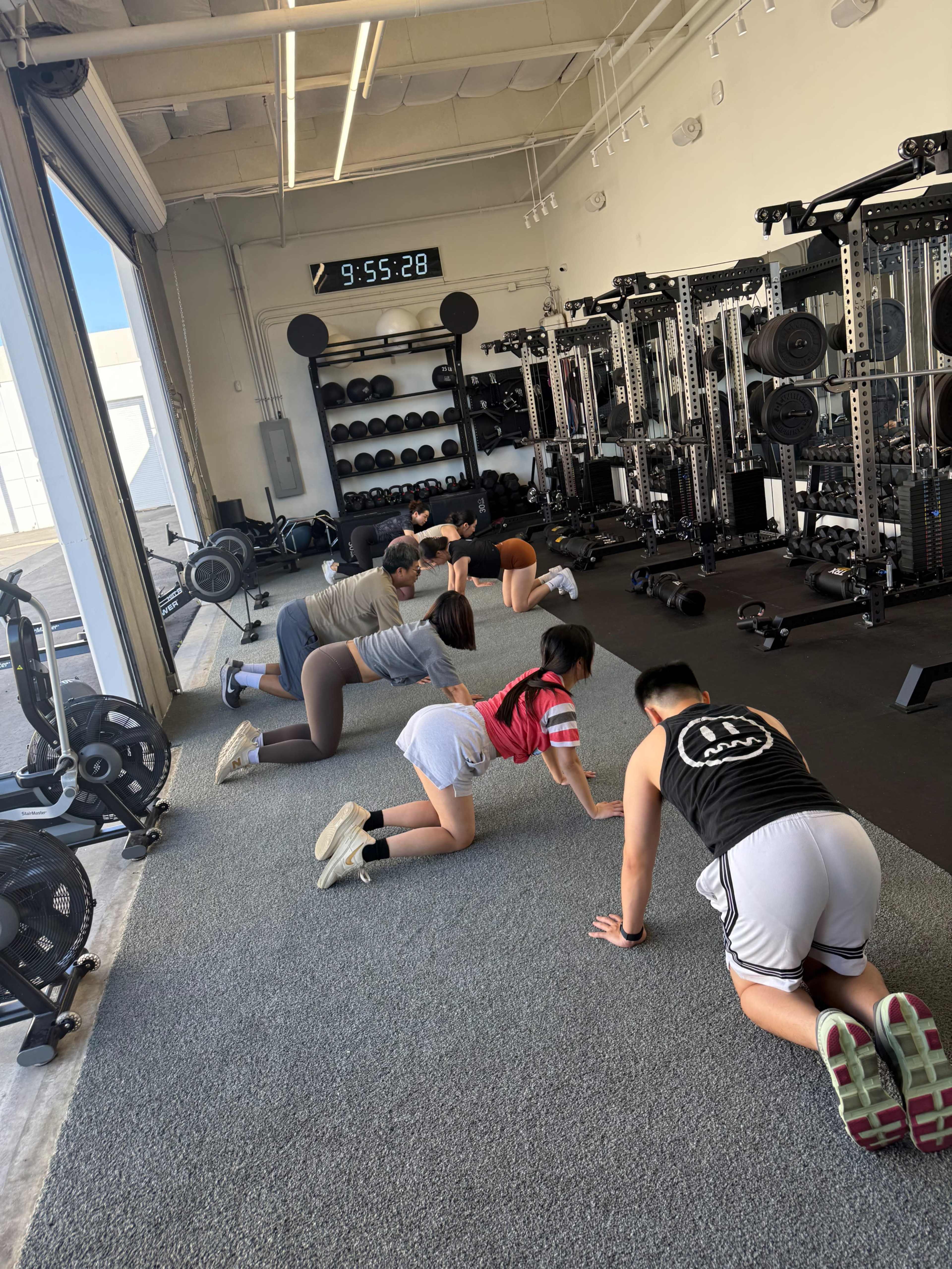 A group of six individuals are exercising on a gray floor in a gym, positioned on all fours with various equipment and weights visible in the background.