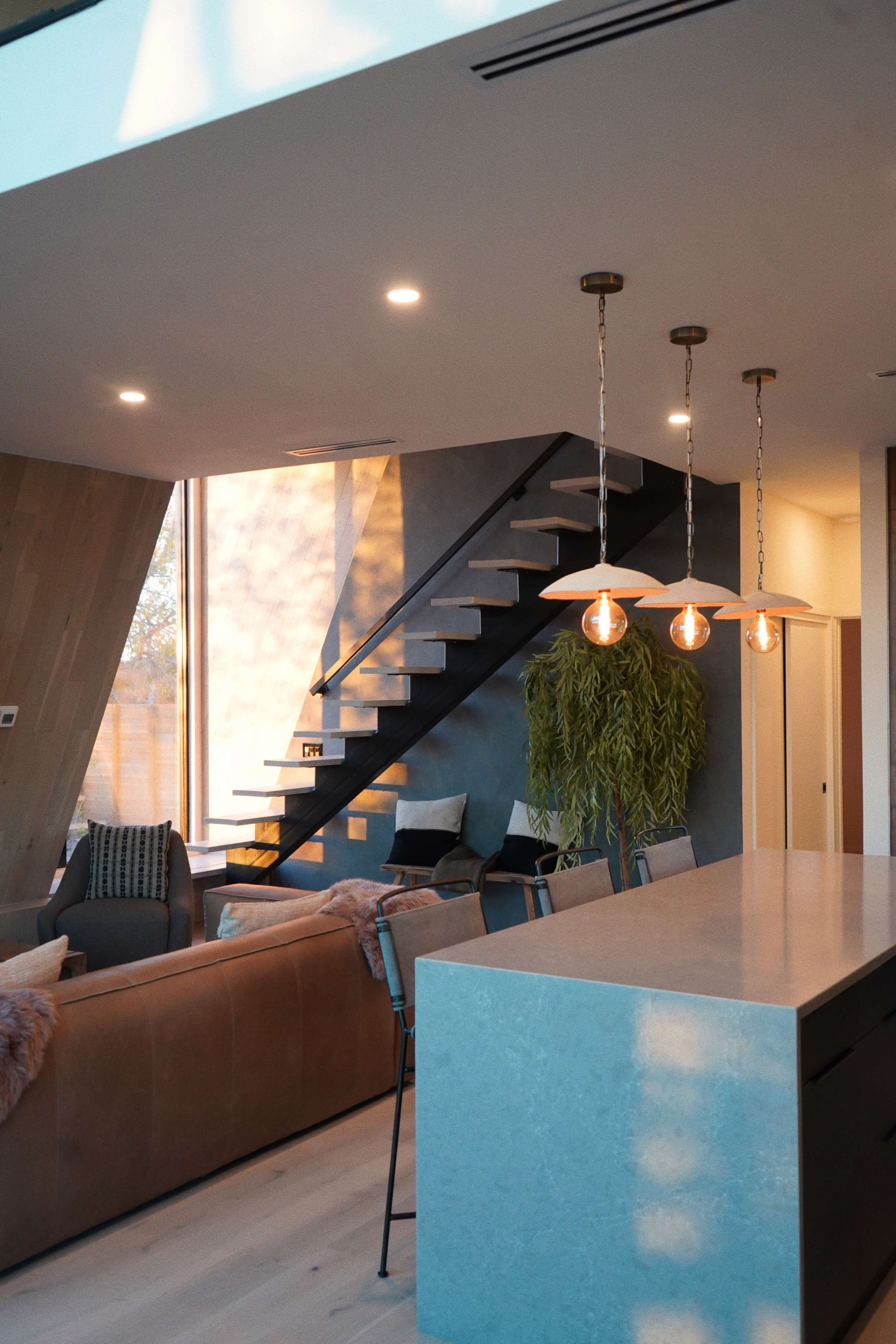 A modern living space featuring a brown leather sofa, a sleek kitchen island with blue countertops, and a staircase with a black railing in a well-lit room.