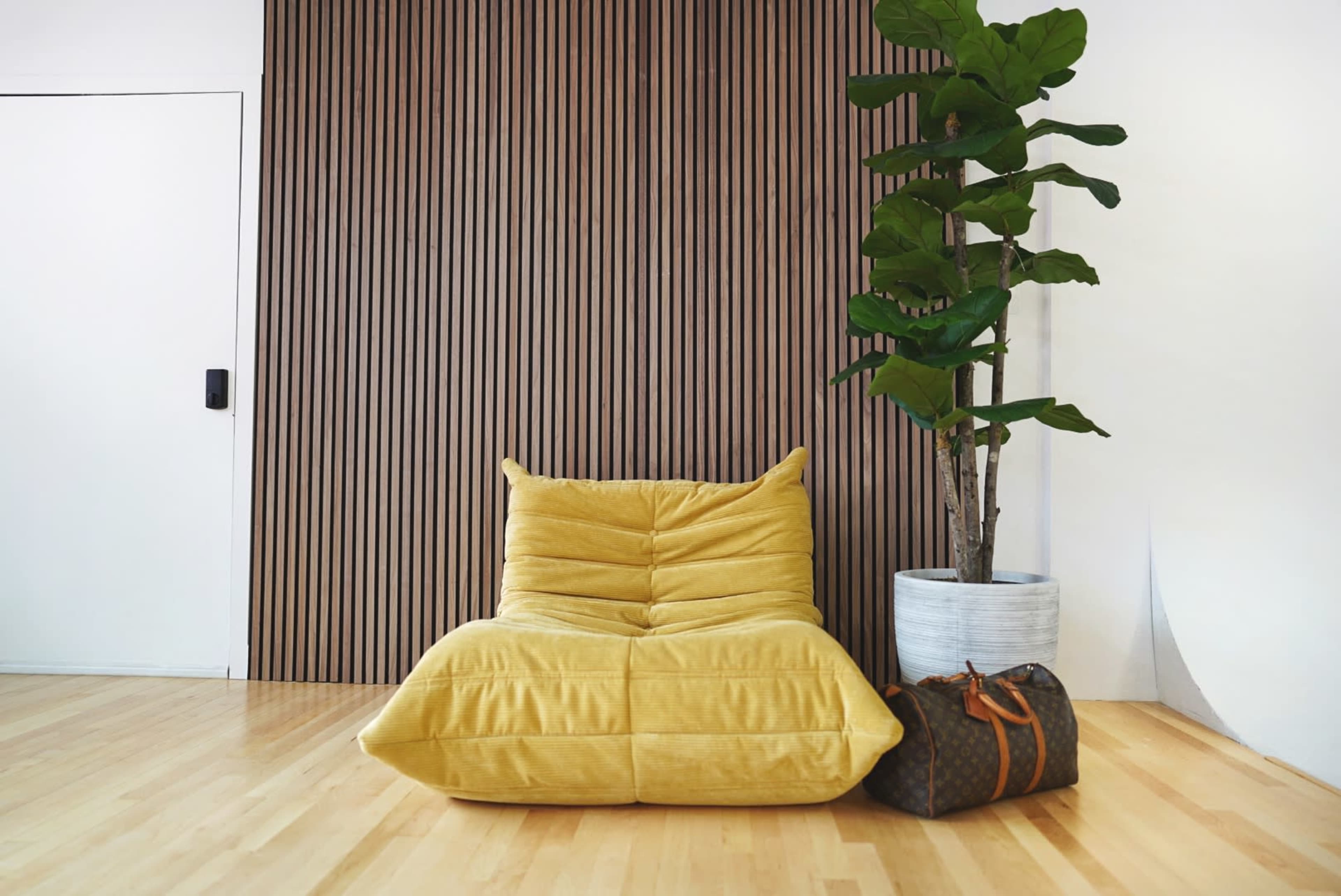 A yellow lounge chair and a potted plant are positioned next to a brown-striped wall and a closed white door in a hardwood-floored room.