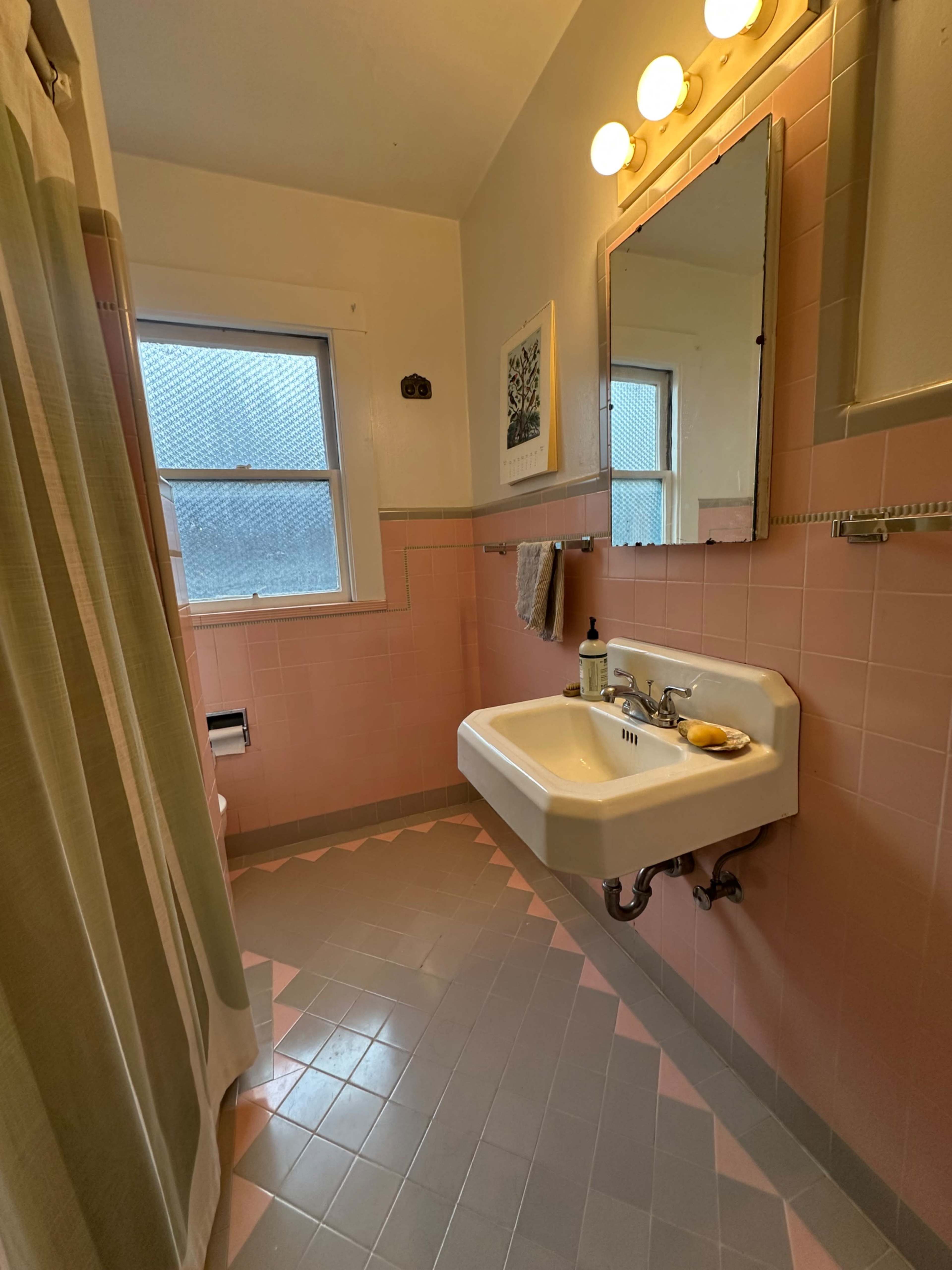 The image shows a bathroom with pink tiled walls, a white sink mounted on a wall, and a shower curtain partially closed.