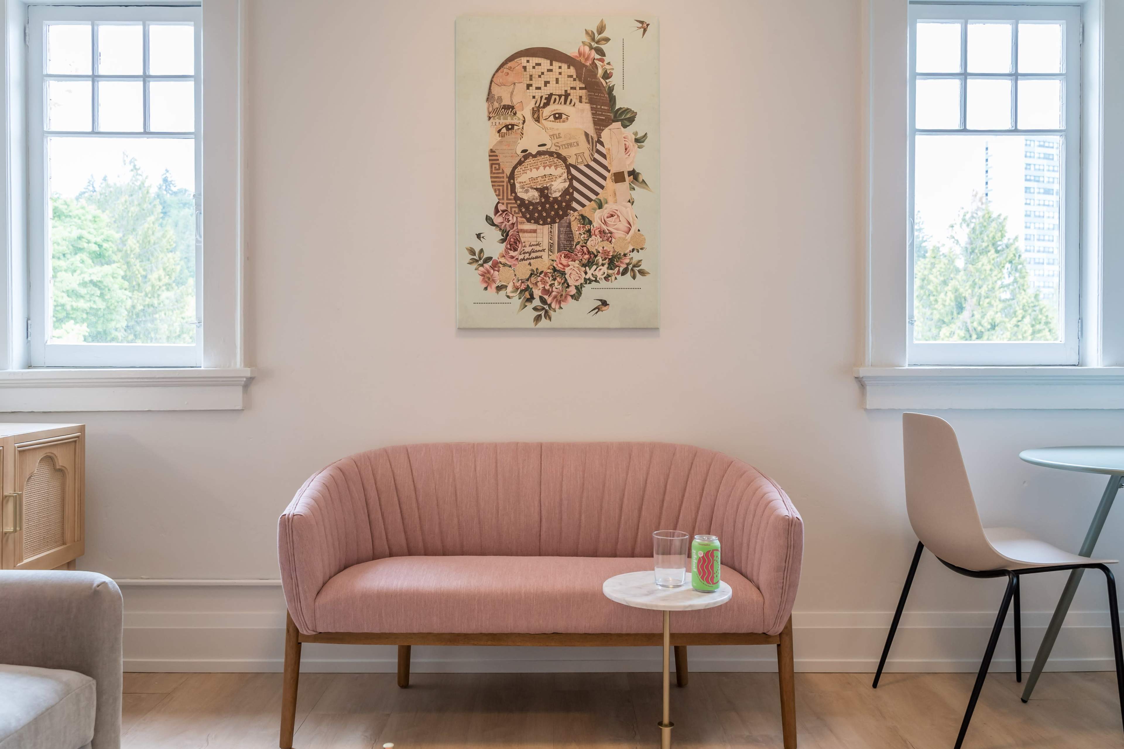 A pink upholstered sofa is positioned against a wall with a floral portrait hanging above it, flanked by two windows, while a small white side table holds a glass and a can, next to a light-colored chair and a round table.