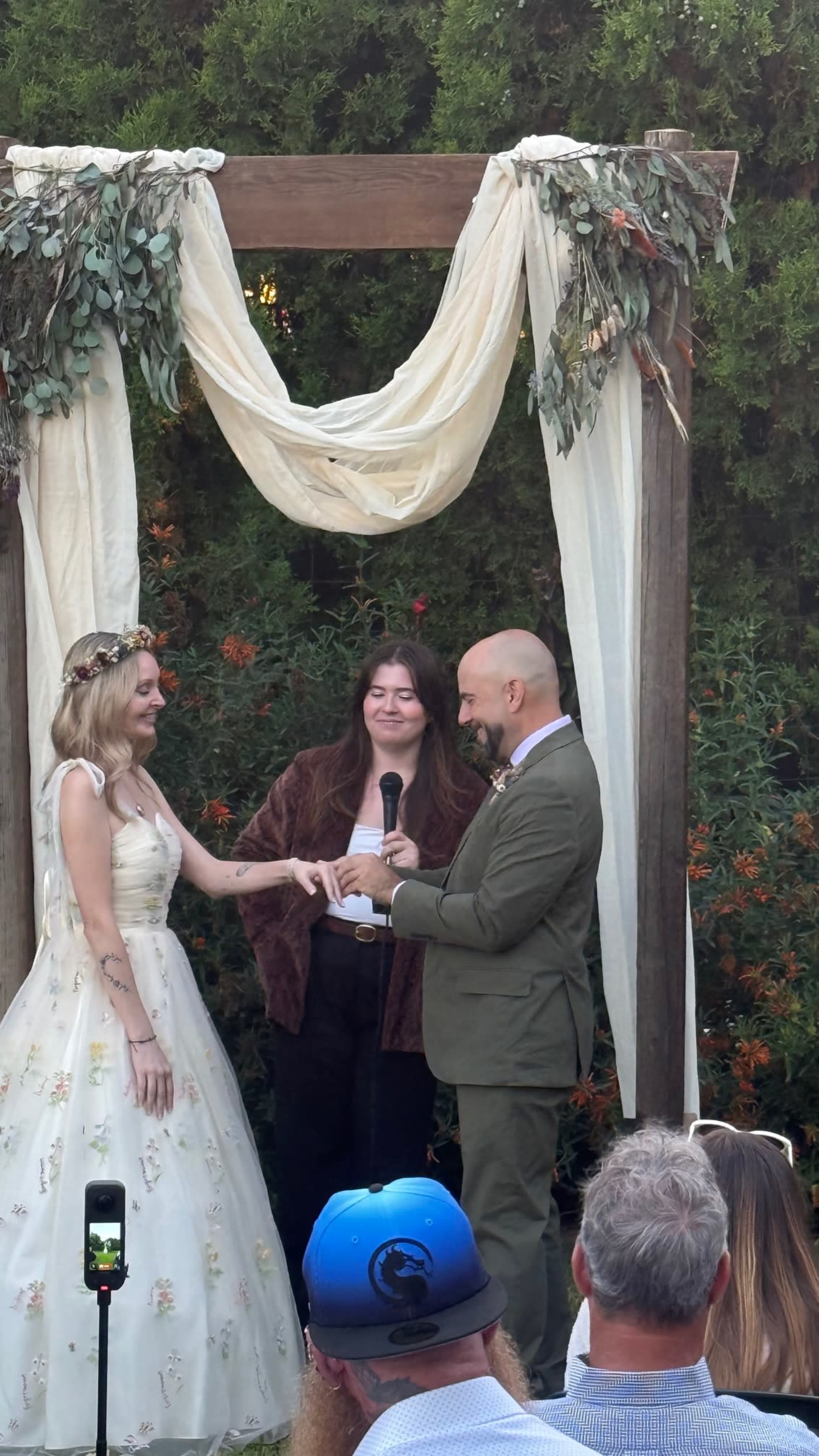 A couple stands under a decorated archway during their wedding ceremony, exchanging vows as guests look on.