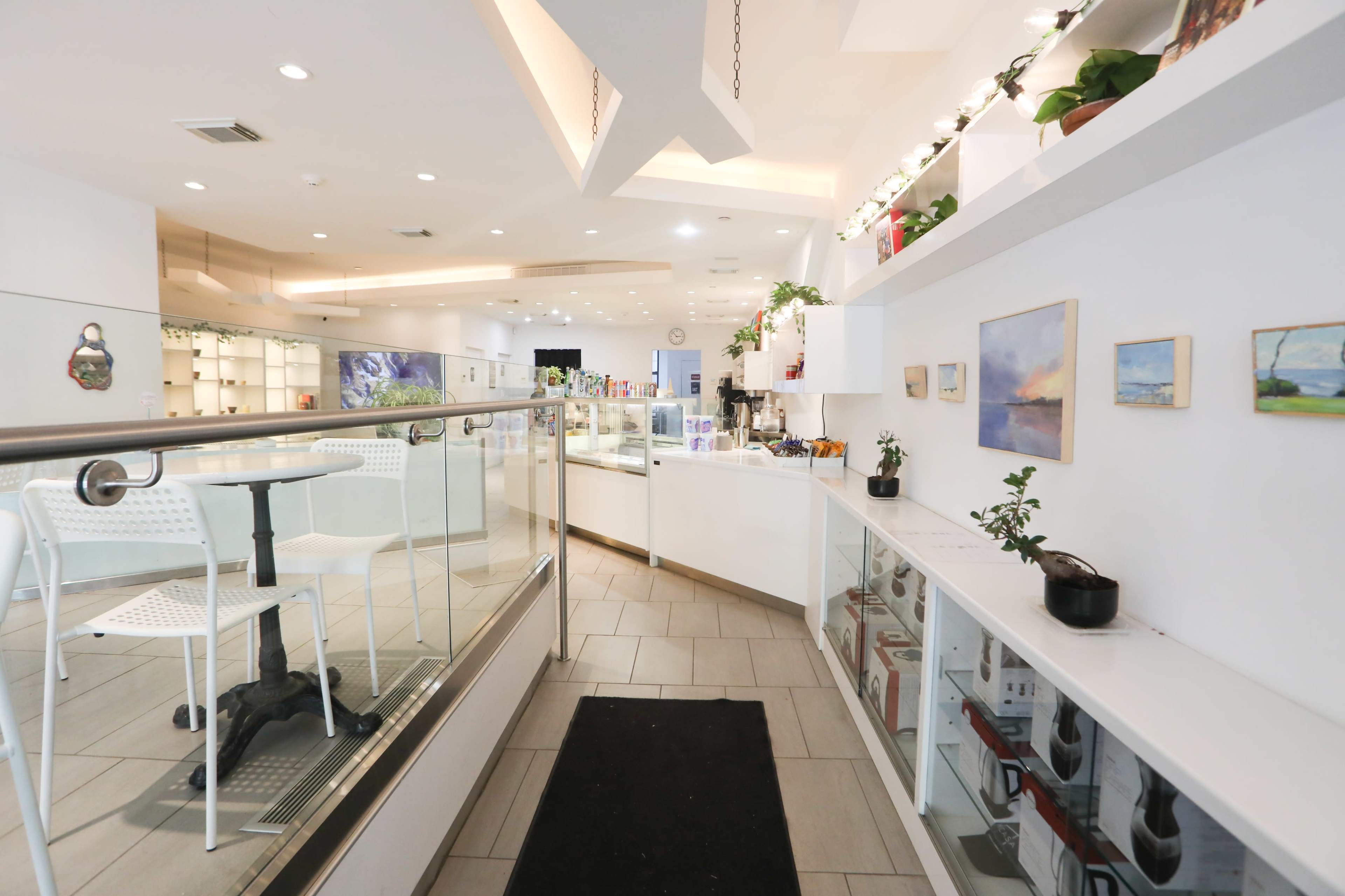 A white-themed café features a display counter, seating area, and decorative plants along the walls.