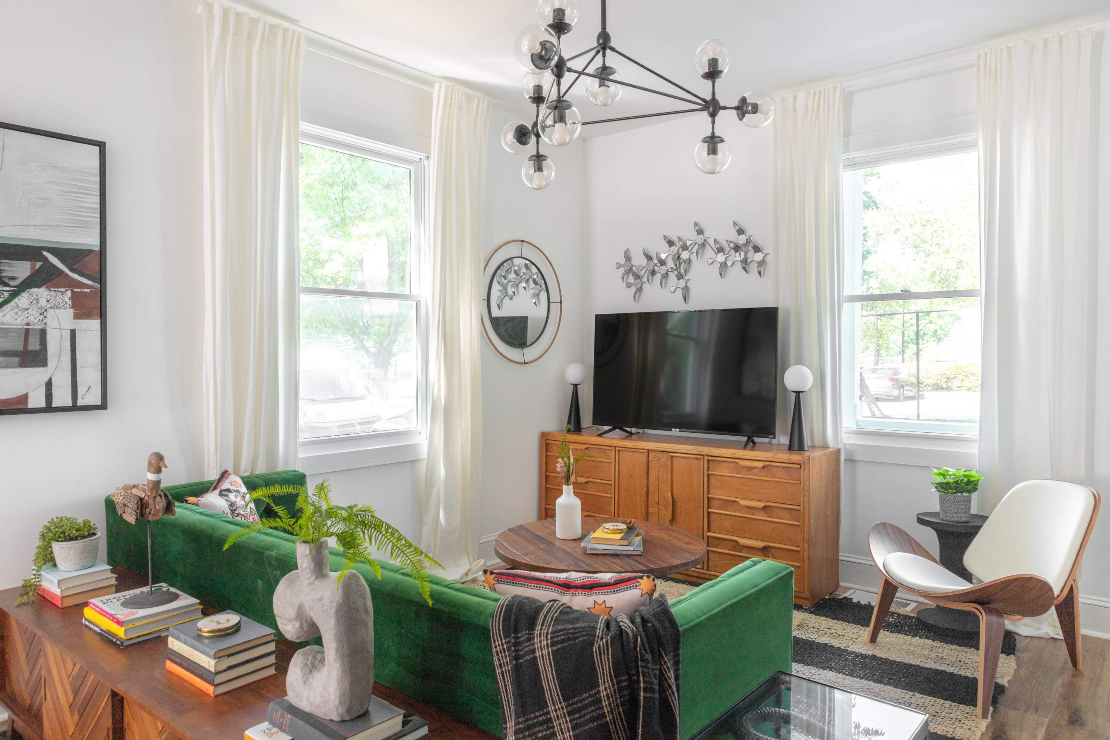 A bright living room featuring a green sofa, a wooden TV cabinet, and large windows with white curtains.