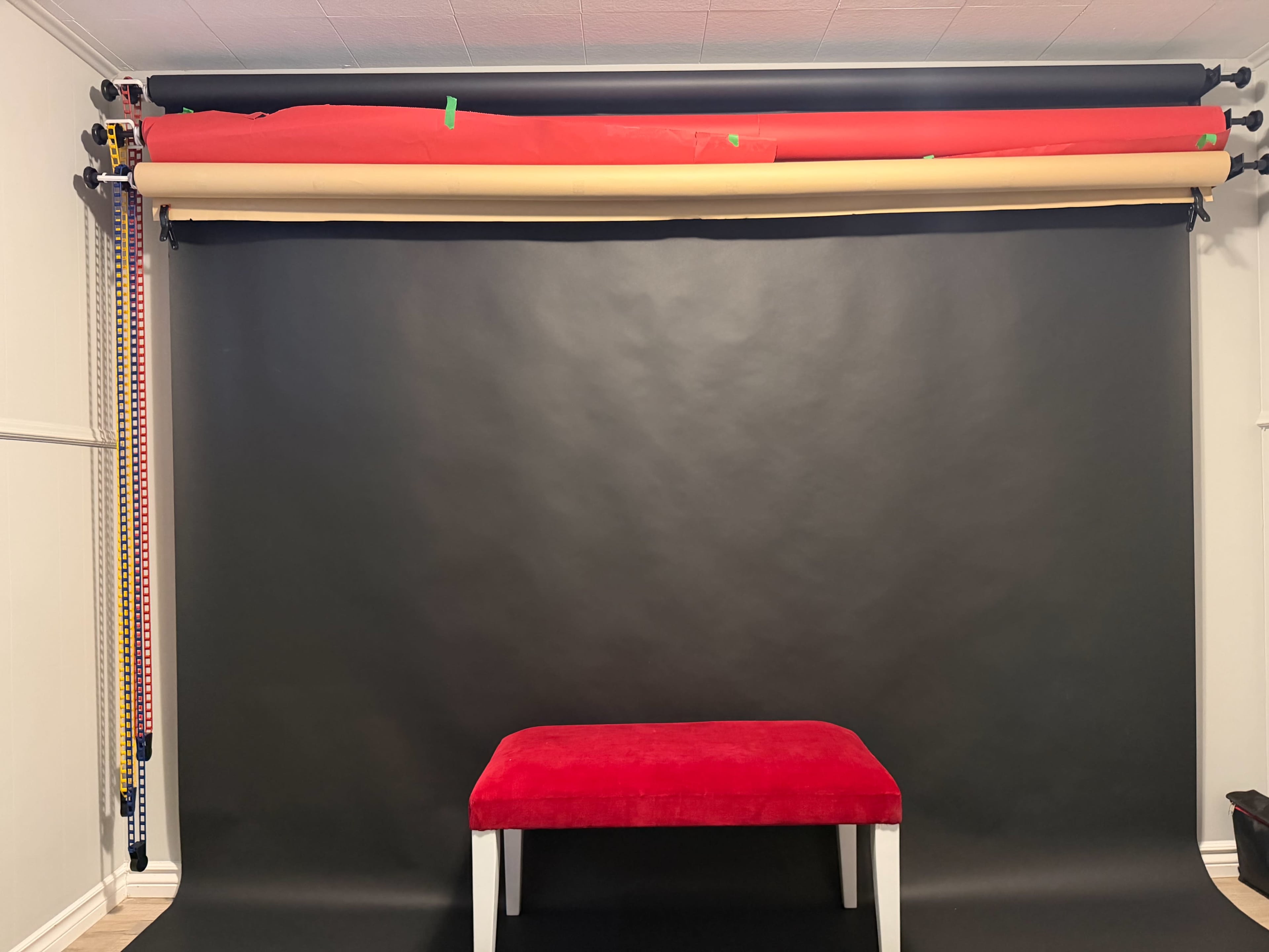 The image shows a photography studio setup with a black backdrop, several rolls of colored paper overhead, and a red velvet bench in front.