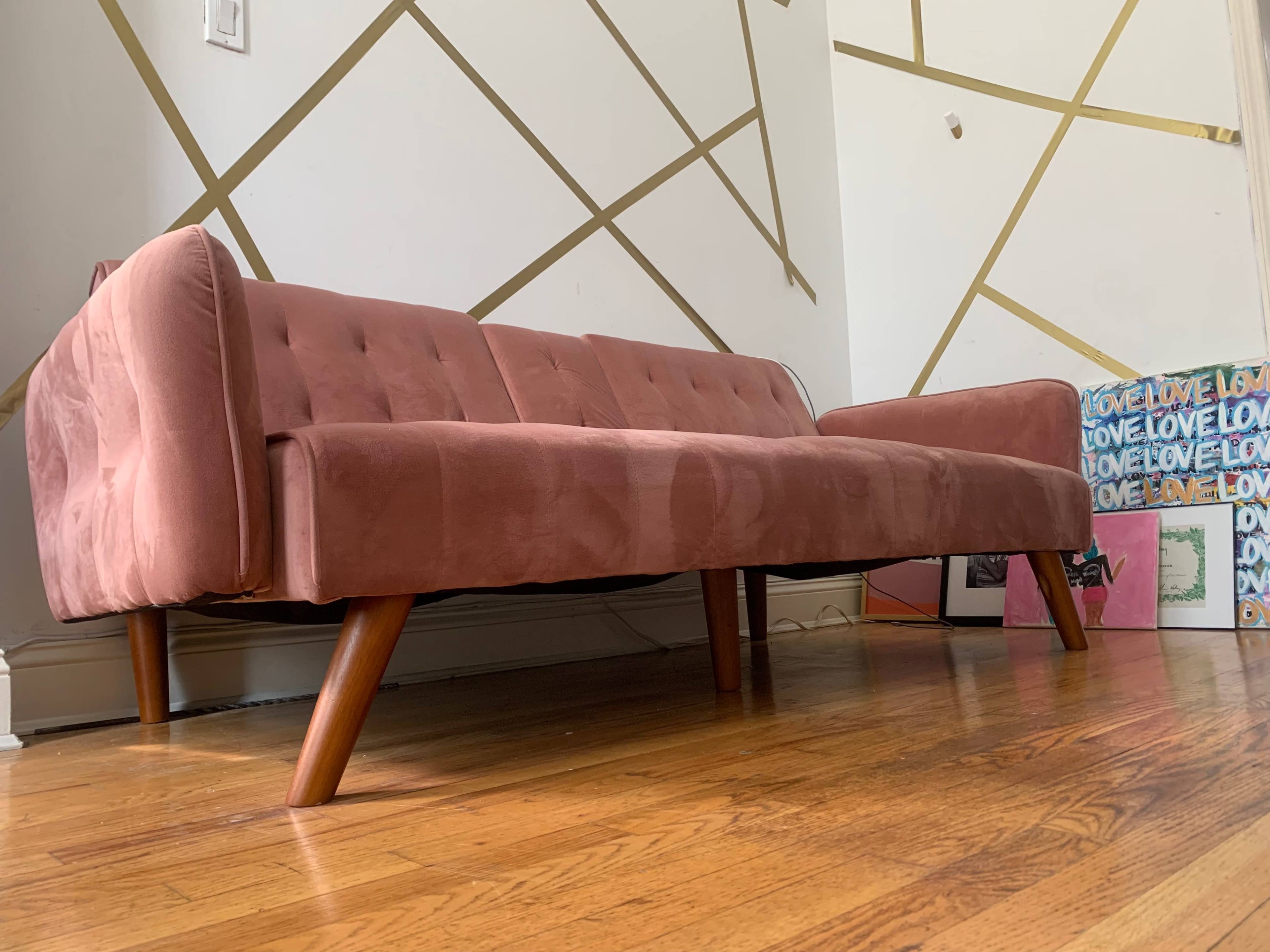 A pink fabric sofa with wooden legs is positioned on a wooden floor against a wall featuring gold geometric designs.