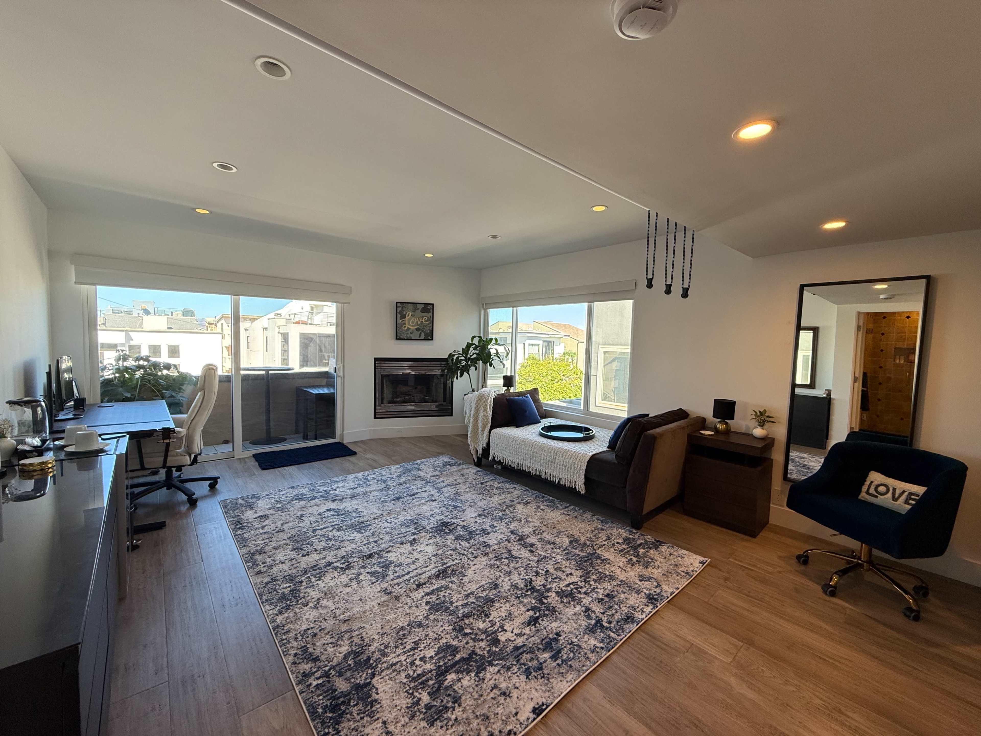 The room features a living area with a sofa, a coffee table, a fireplace, large windows, and a desk setup, all decorated with a rug and plants.