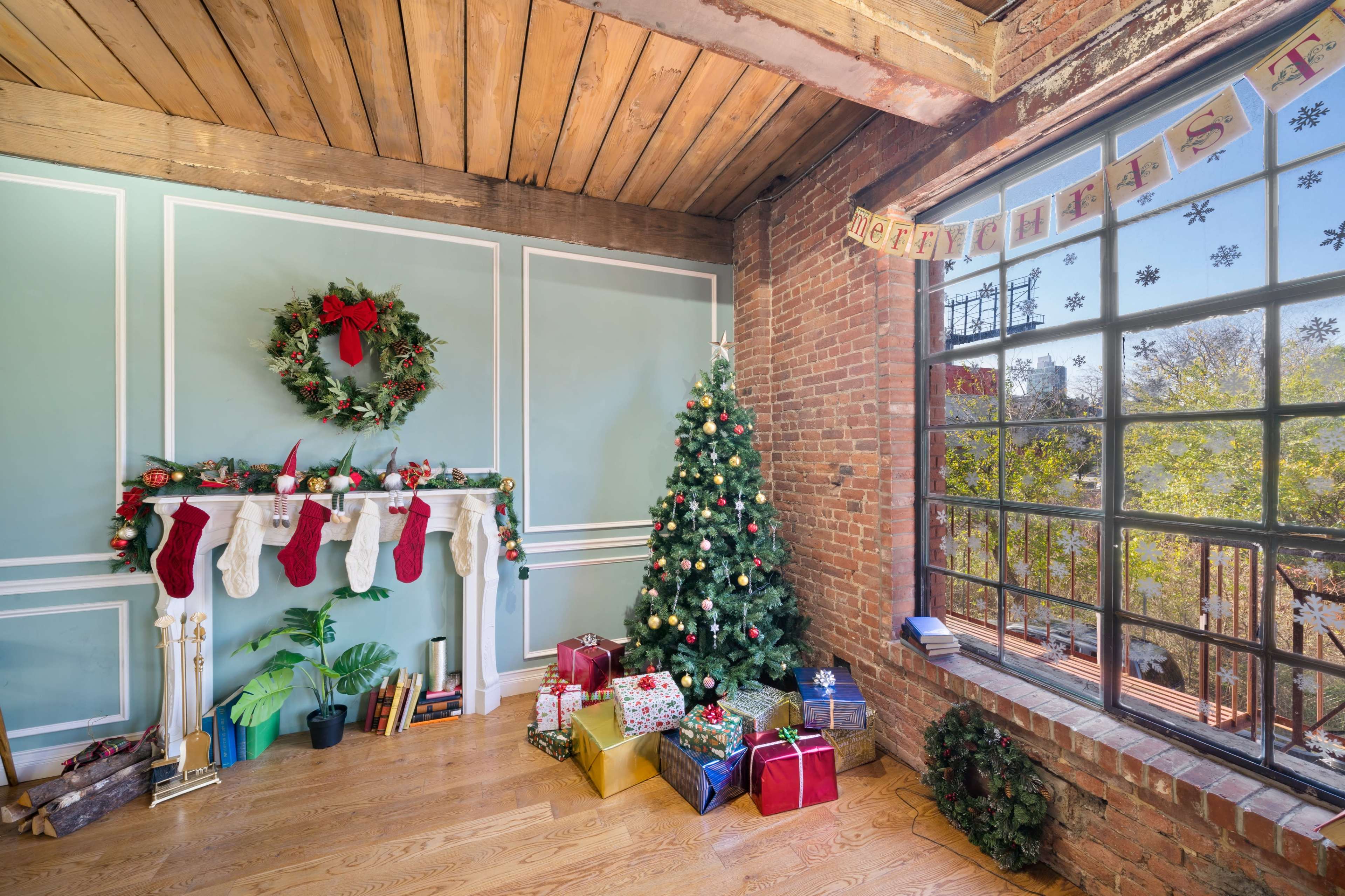 A cozy room features a Christmas tree, stockings hanging from a mantel, and gifts arranged around it, with large windows showcasing the outside.