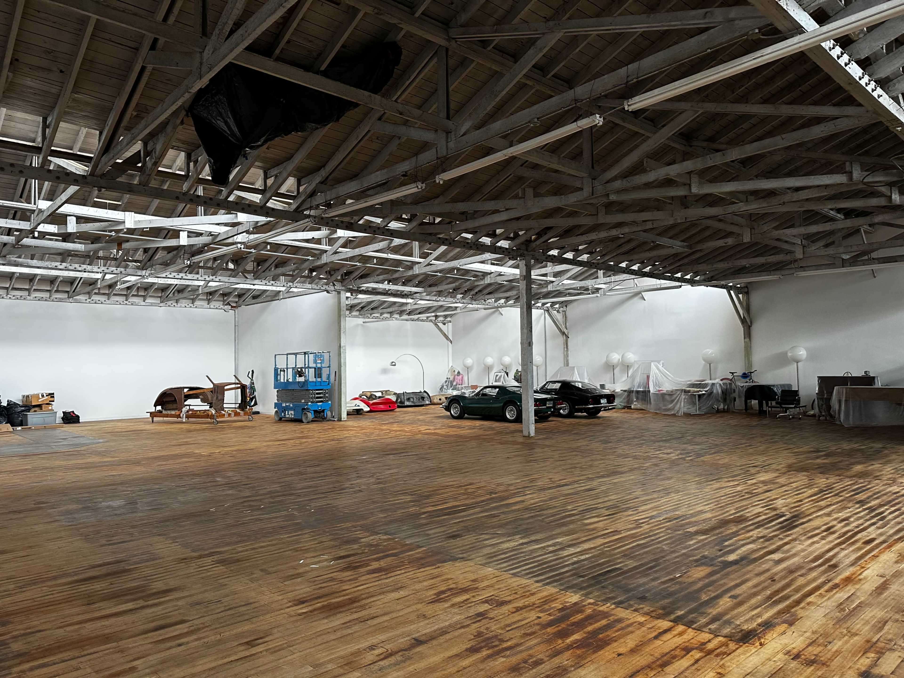 A spacious industrial-style warehouse with wooden flooring and several cars, including a classic model, in the background.