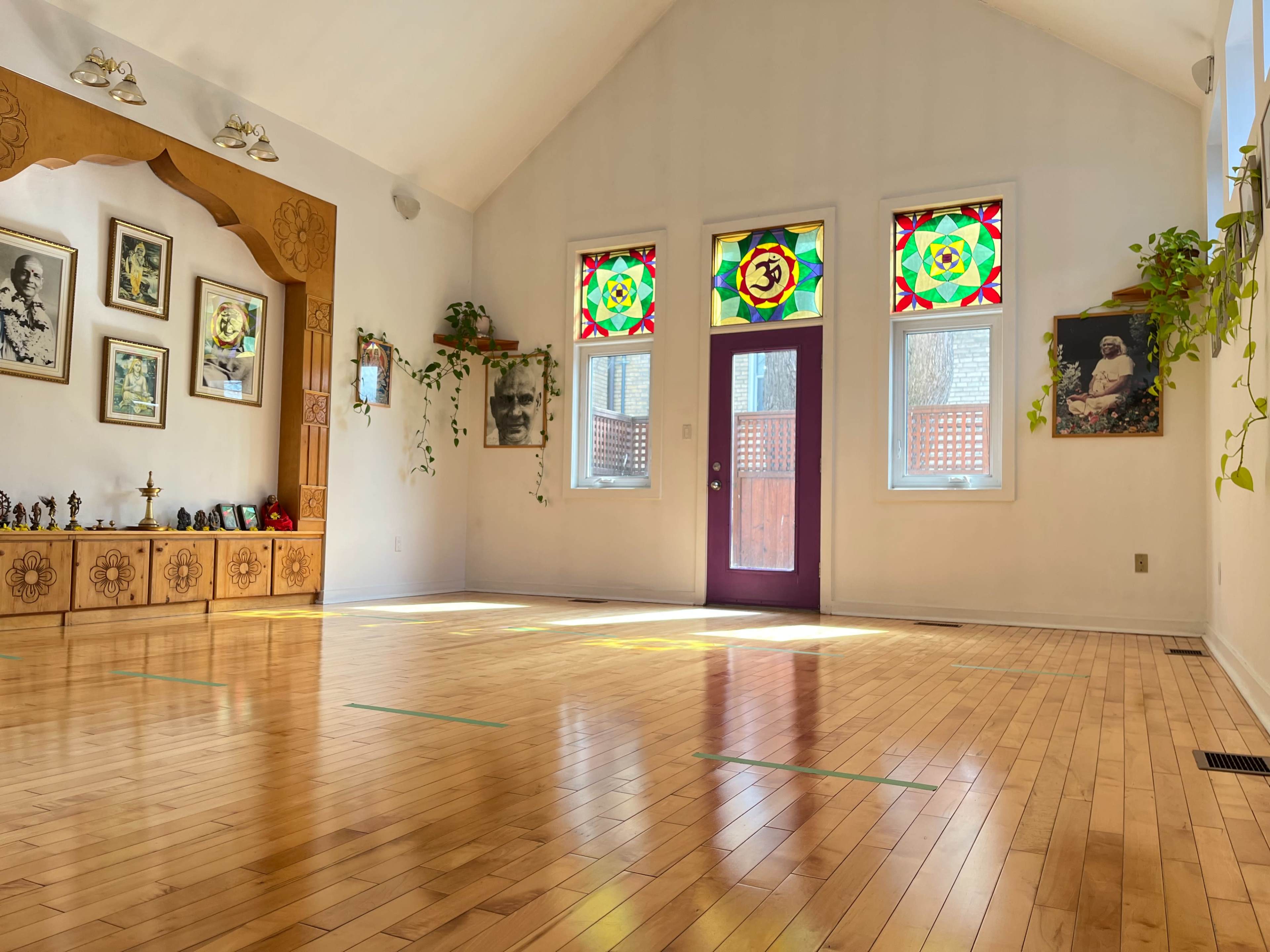 A spacious, well-lit room with wooden flooring, featuring colorful stained glass windows and a decorative wooden wall with framed photographs.