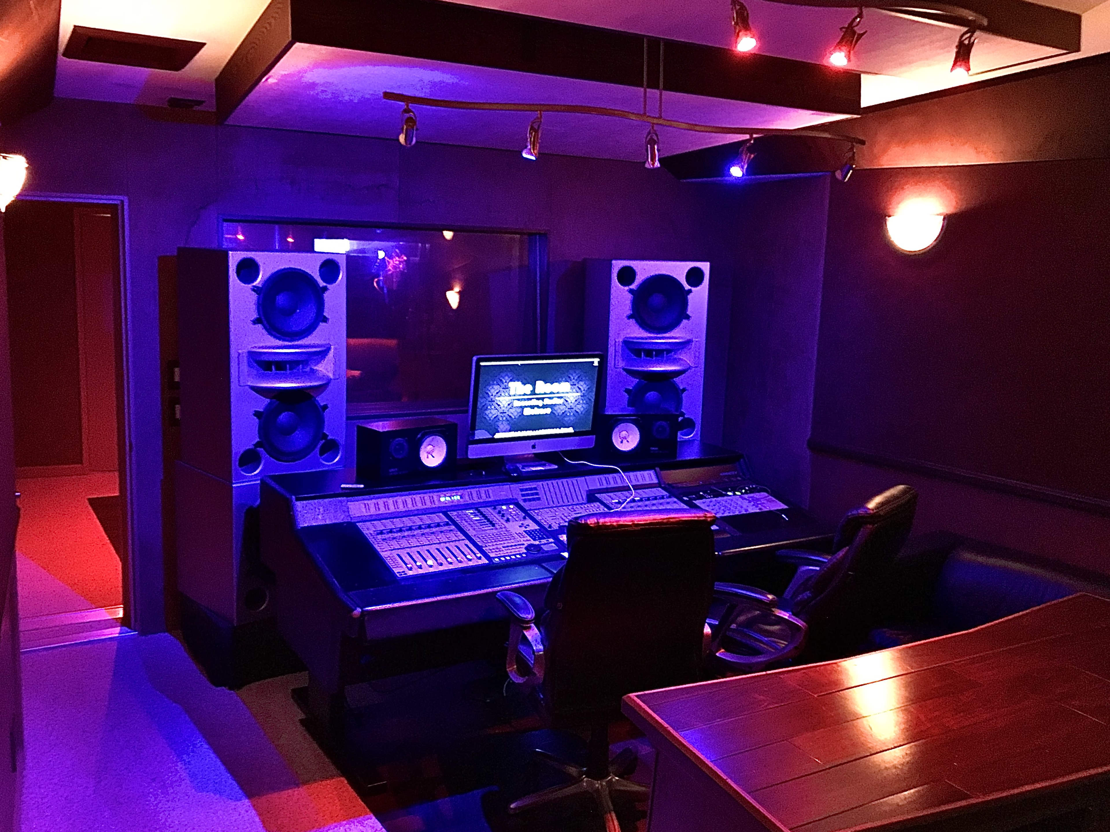 The image shows a music recording studio featuring a control desk with a large computer monitor, speakers, and dim lighting in various shades.