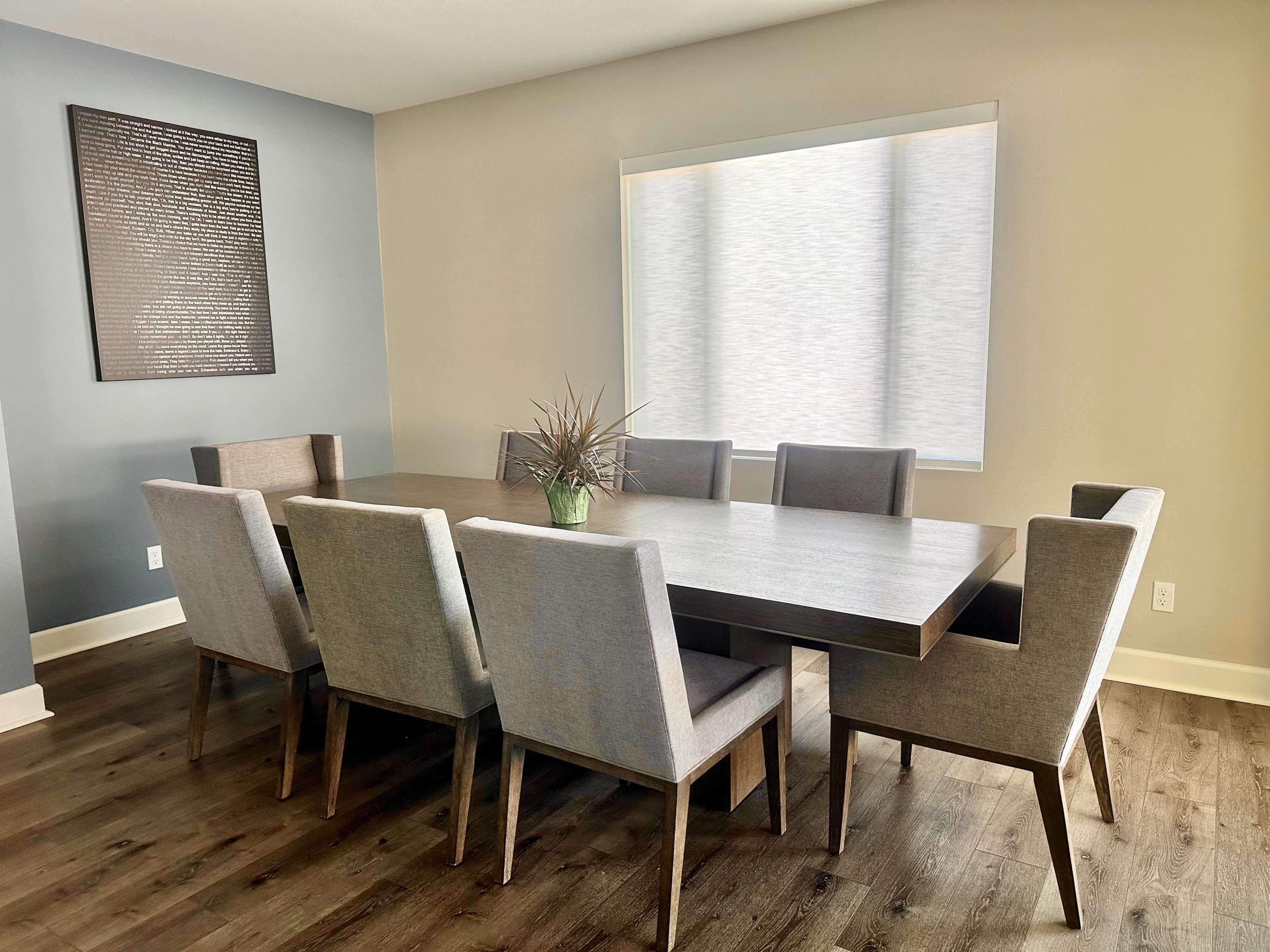 A modern dining room features a large wooden table surrounded by eight upholstered chairs and a potted plant in the center.