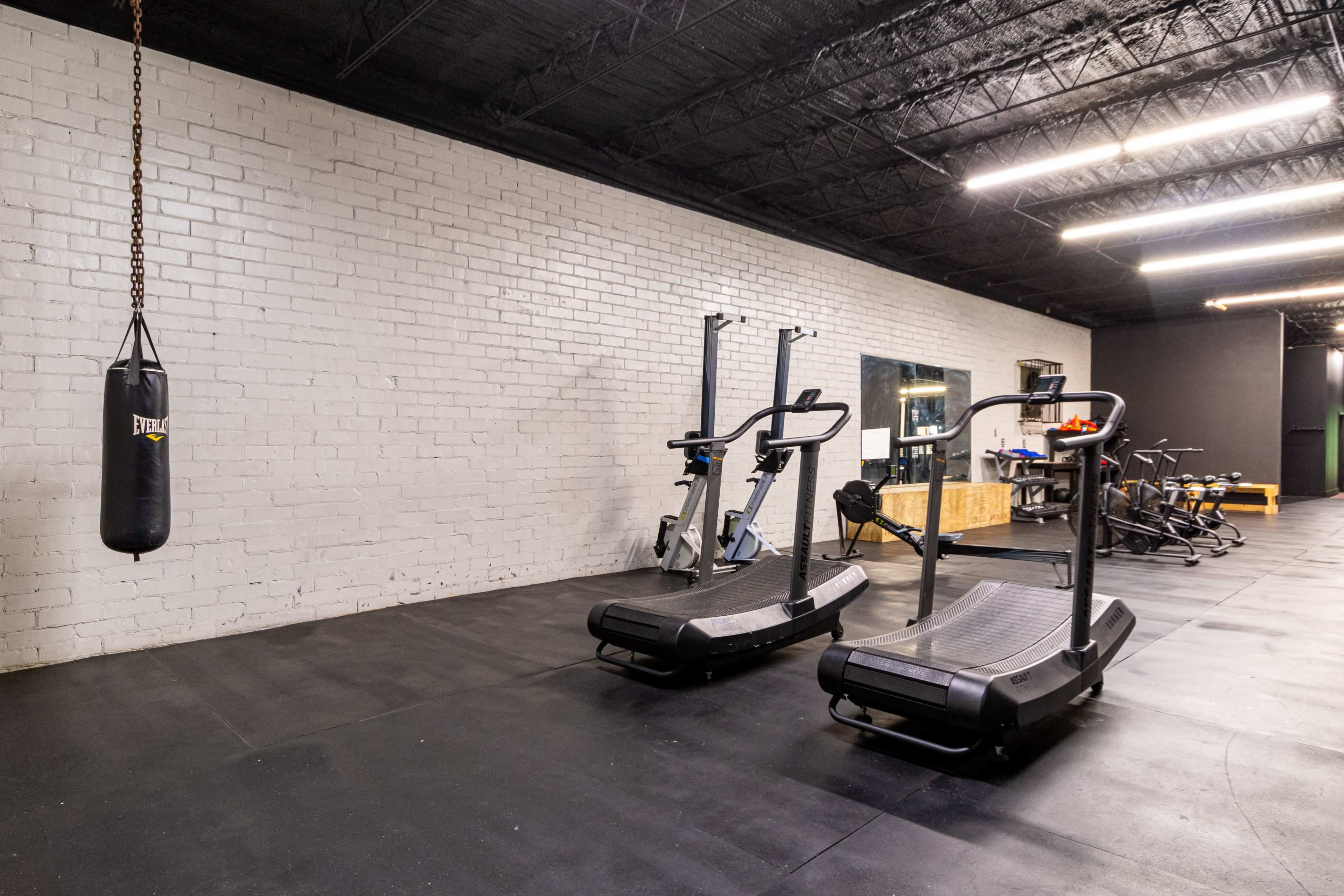 A gym interior featuring two treadmills, a punching bag hanging from the ceiling, and various exercise equipment against a white brick wall.
